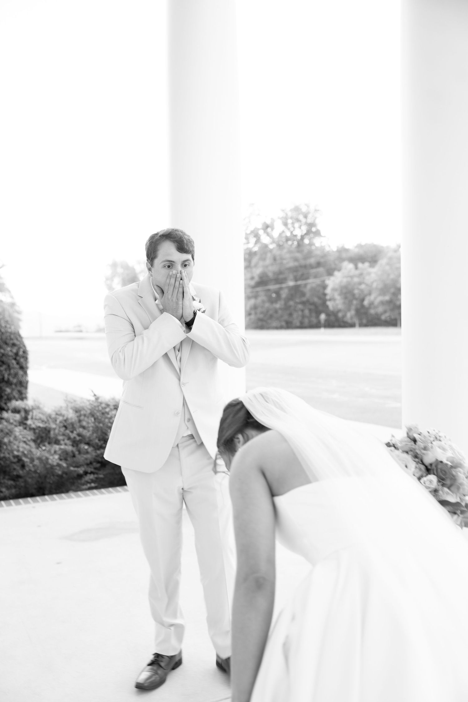 Emotional groom covers his mouth in awe as bride bows during their first look at a wedding venue.