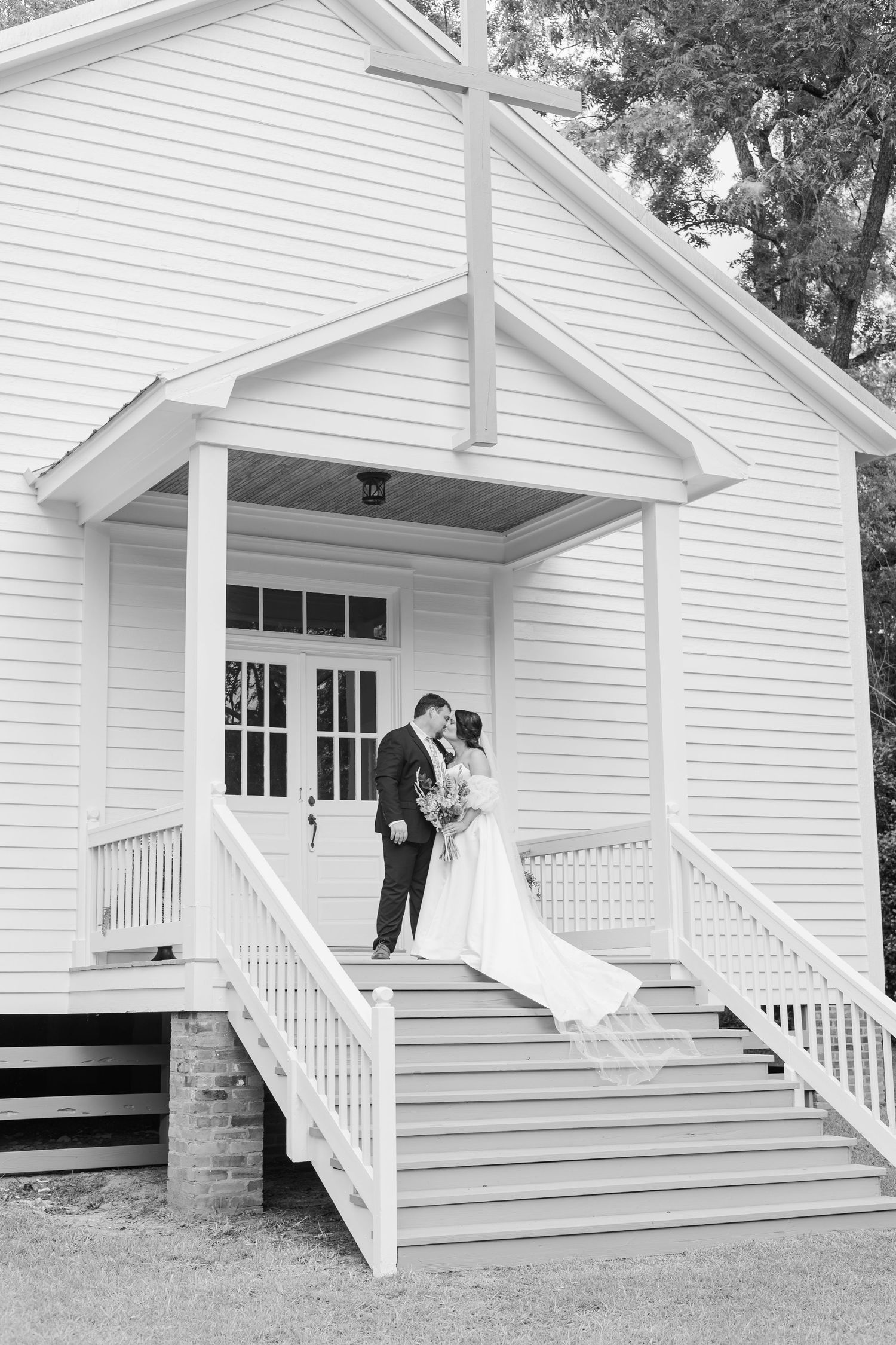 Bride and groom share a kiss on steps of charming white chapel with cross in classic black and white wedding photo.