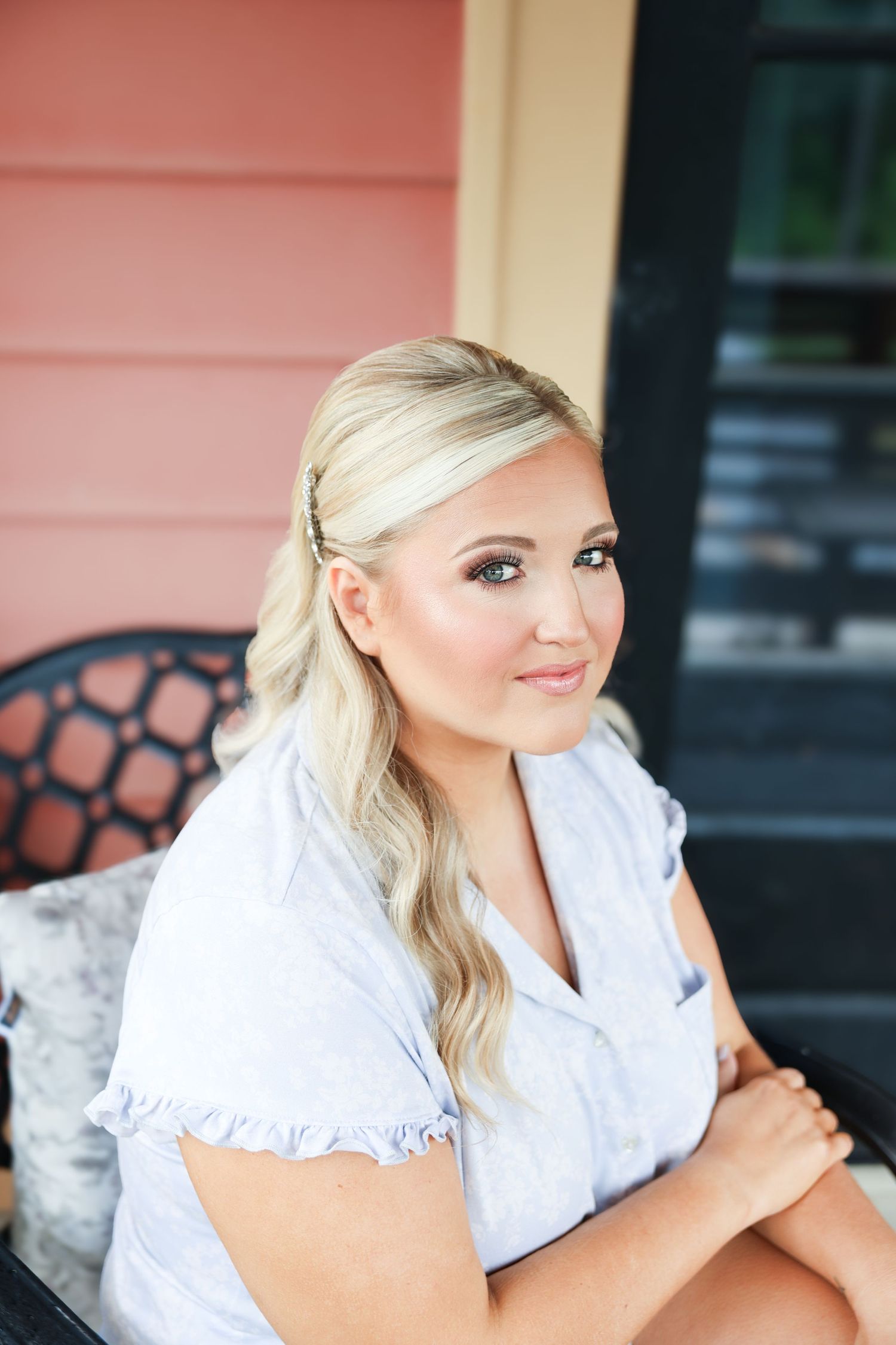 A blonde woman in a light blue ruffled top sits outdoors smiling, with a pink building in the background.