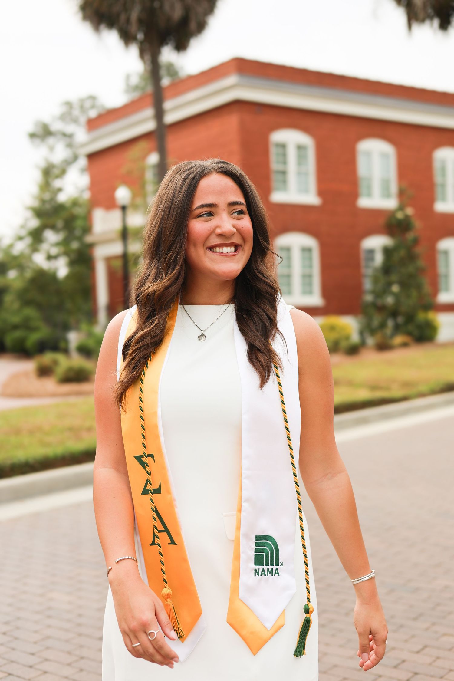 Graduate in white dress and gold stole poses in front of red brick building during commencement photo session.
