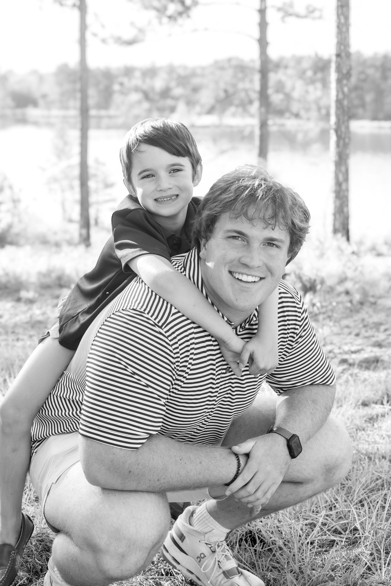 Black and white photo of a smiling man crouching outdoors with a young boy on his back near a lake.
