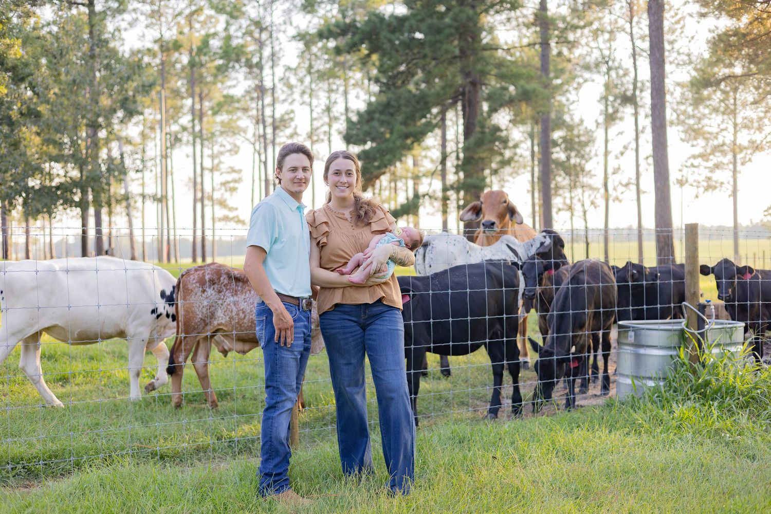 Family of three with baby posing by cattle fence on a green farm with trees in background.
