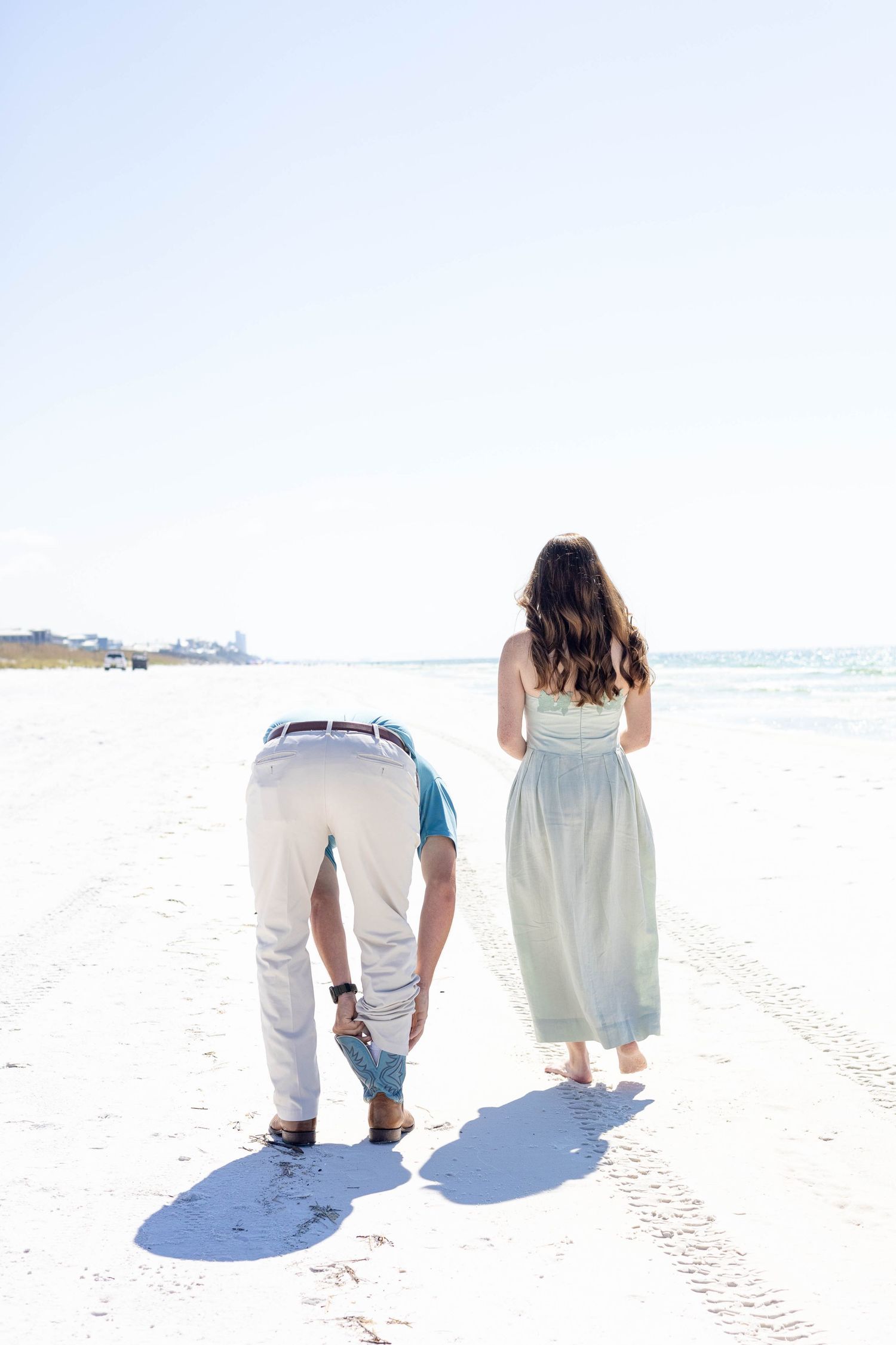 Couple walking on a white sand beach, man bending down and woman in a light green maxi dress, sunny day.