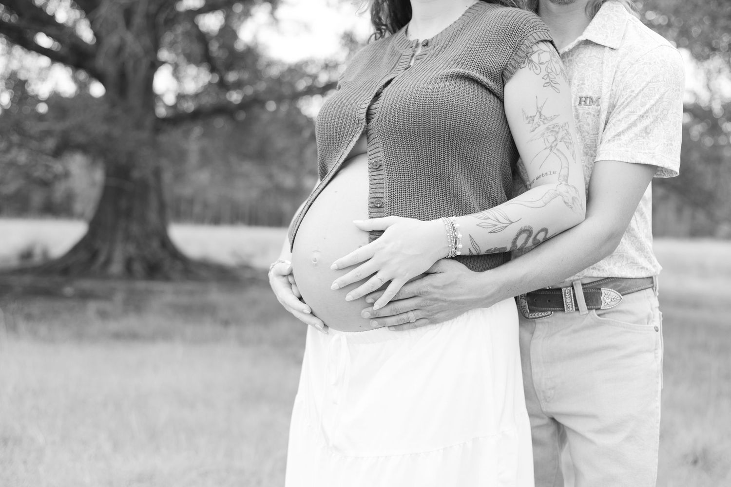 Black and white maternity photo of couple outdoors, man embracing pregnant woman's belly near large tree.