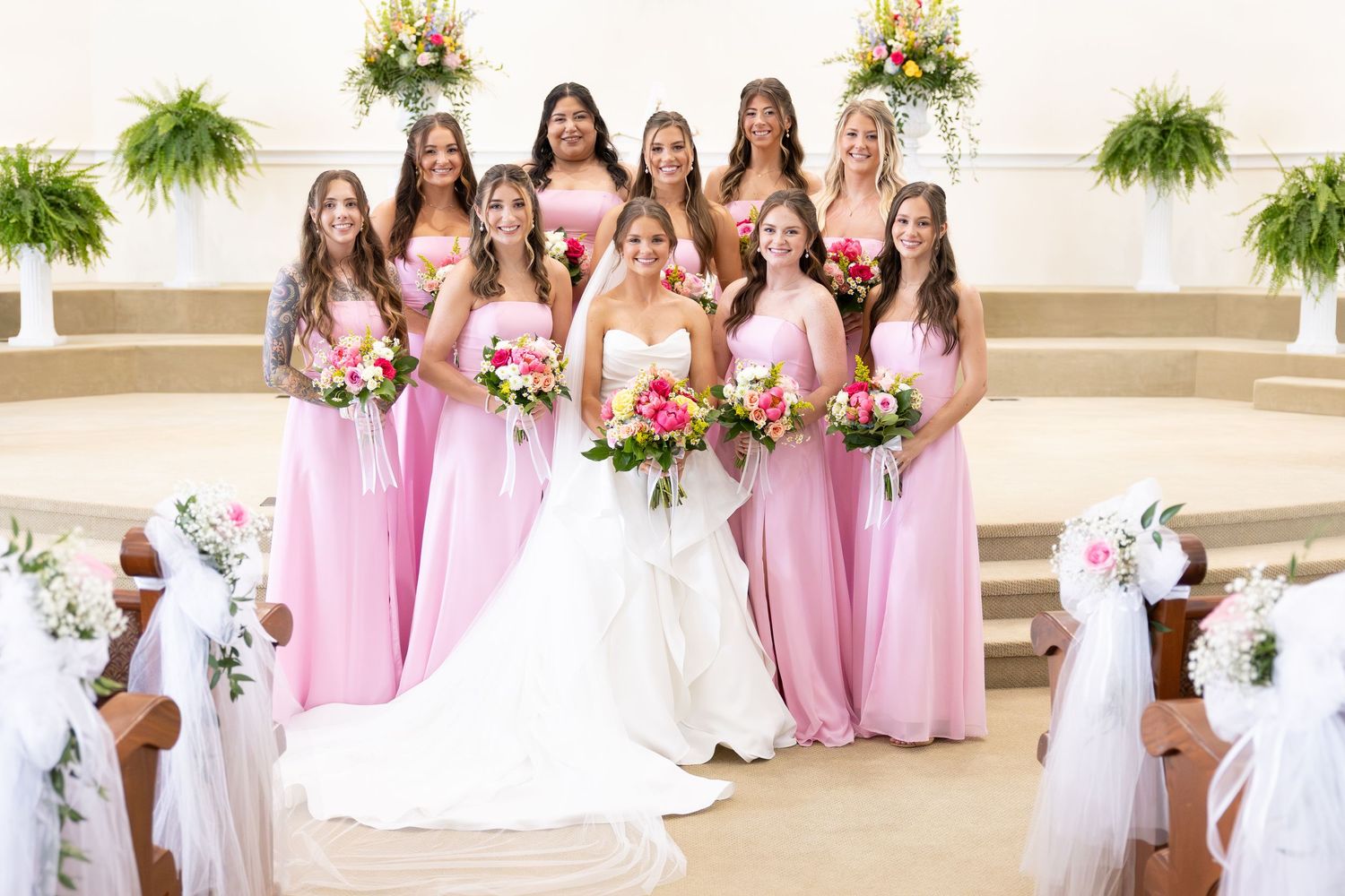 Bride in white gown poses with eight bridesmaids in pink dresses holding bouquets inside a church.