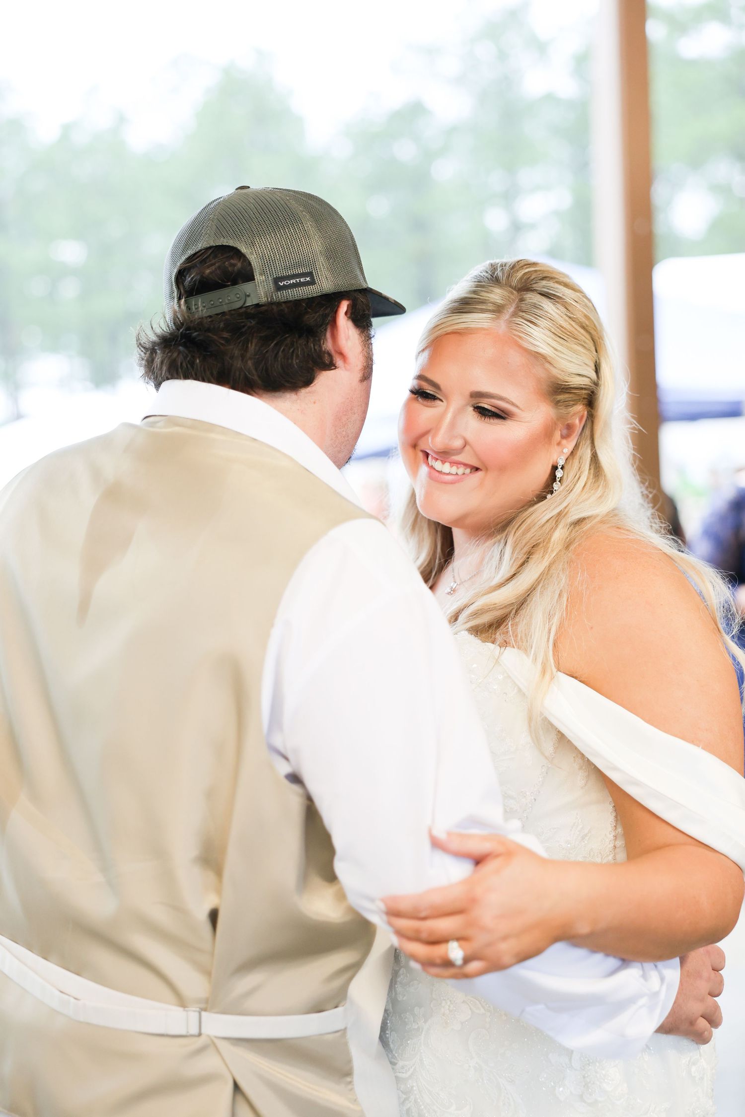 A smiling blonde bride in an off-shoulder gown dances joyfully with a guest in a tan vest and cap.