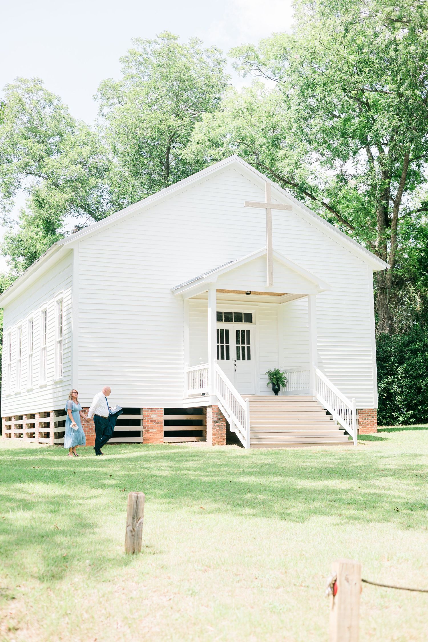 Couple walking outside a white wooden country church with a cross, surrounded by green trees on a sunny day.