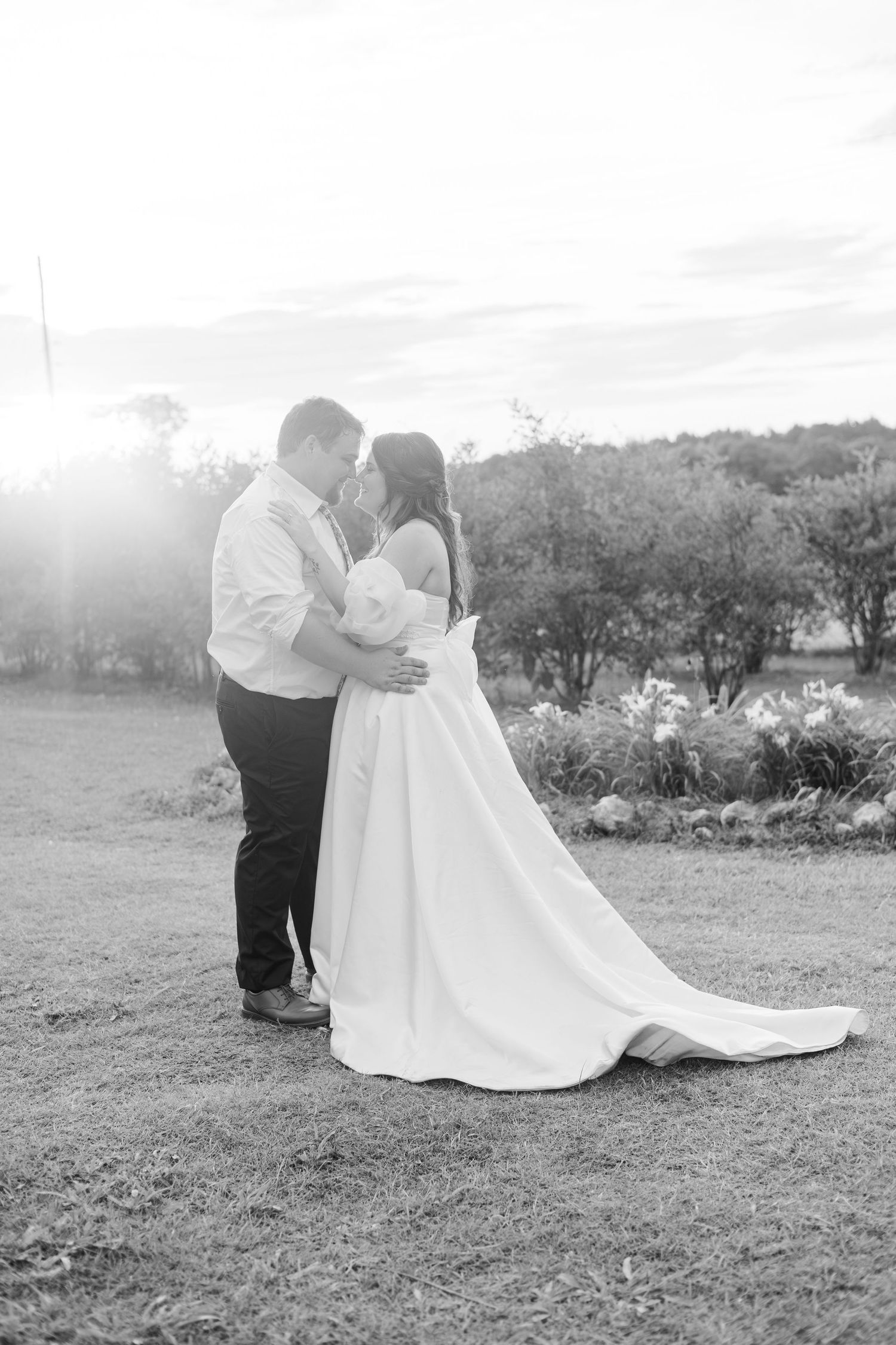 Black and white photo of bride and groom embracing at sunset in garden wedding setting.