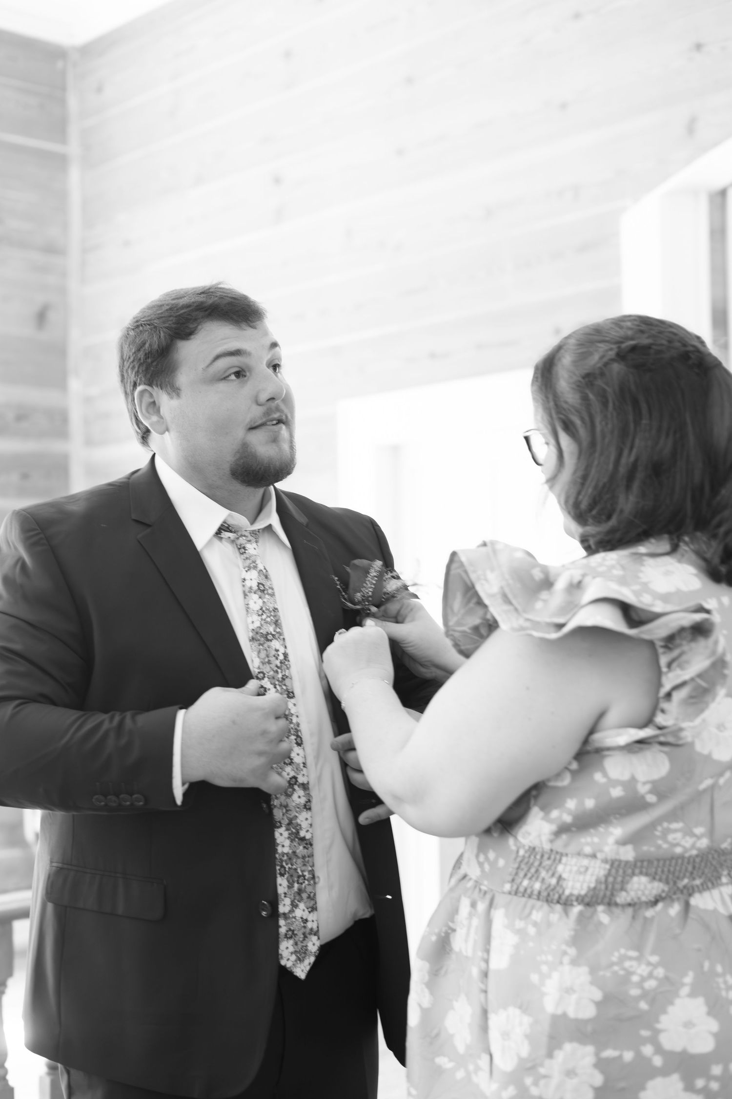 Black and white photo of a woman pinning a boutonniere on a groom in a suit and floral tie.