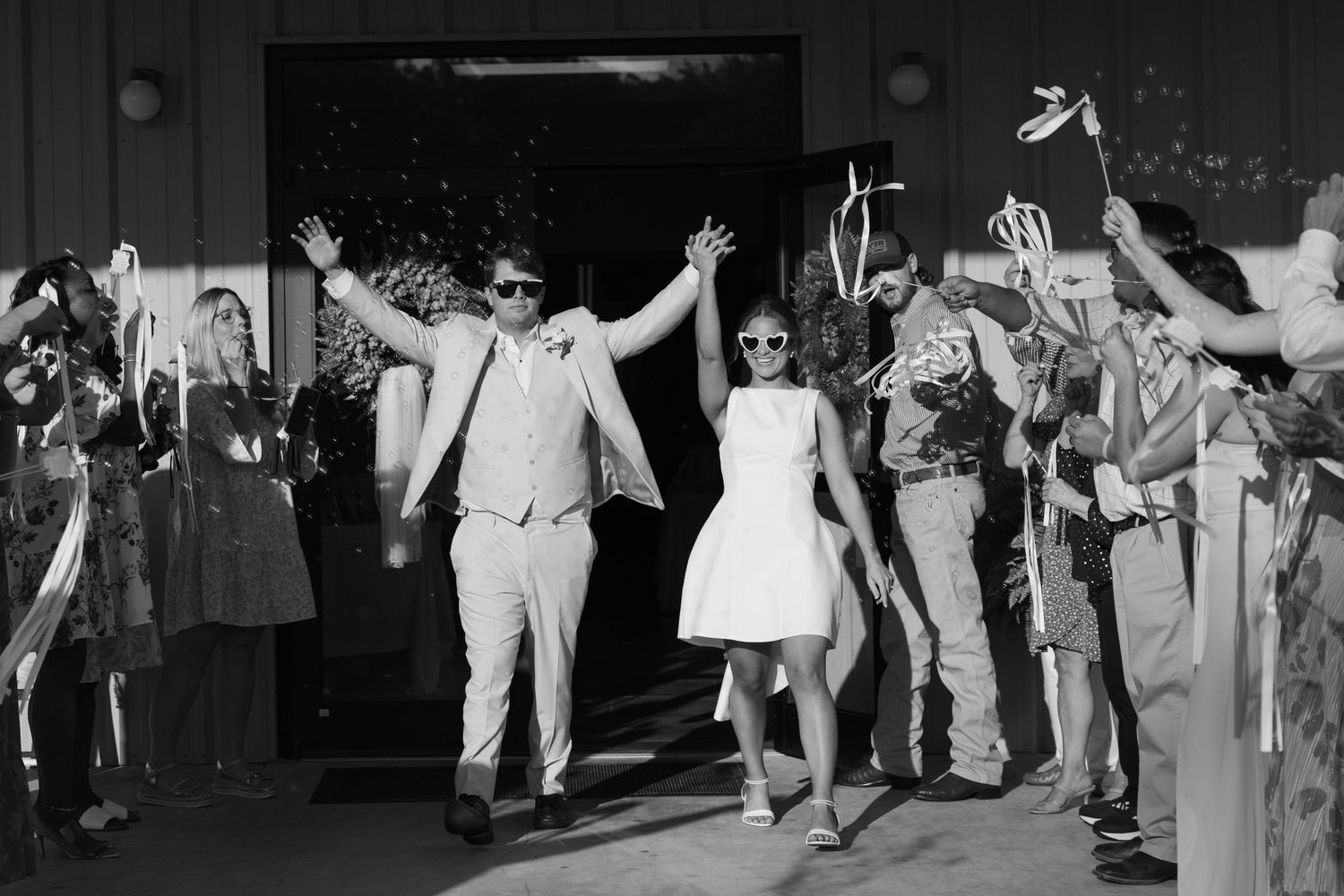 Newlyweds exit wedding venue in sunglasses as guests celebrate with ribbons in joyful black and white photo.