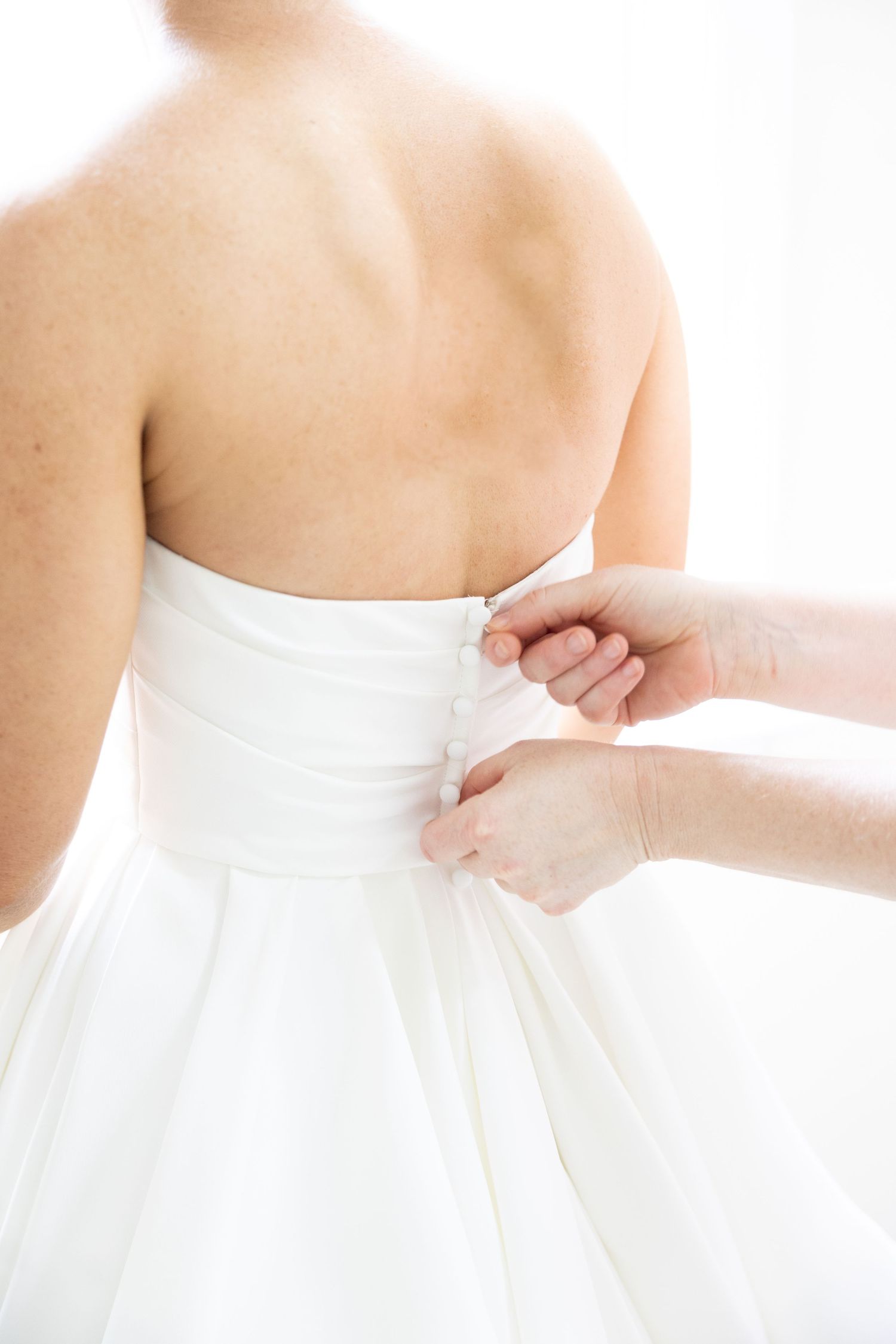 Hands fastening the back zipper of a white strapless wedding dress on a bride.