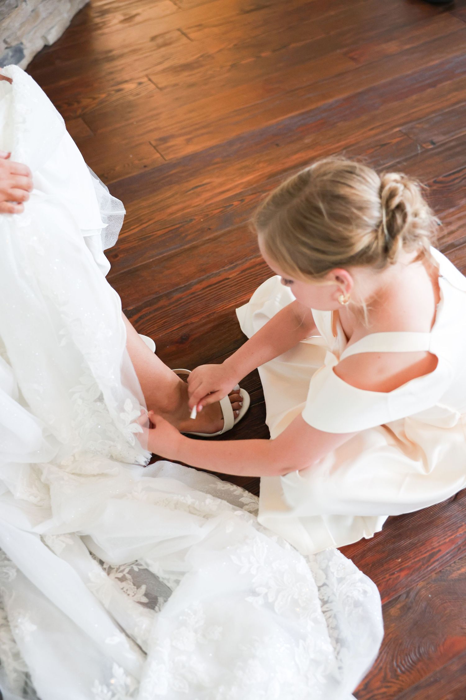 Bridesmaid in ivory dress helping bride with white heeled sandal on hardwood floor during wedding preparation.