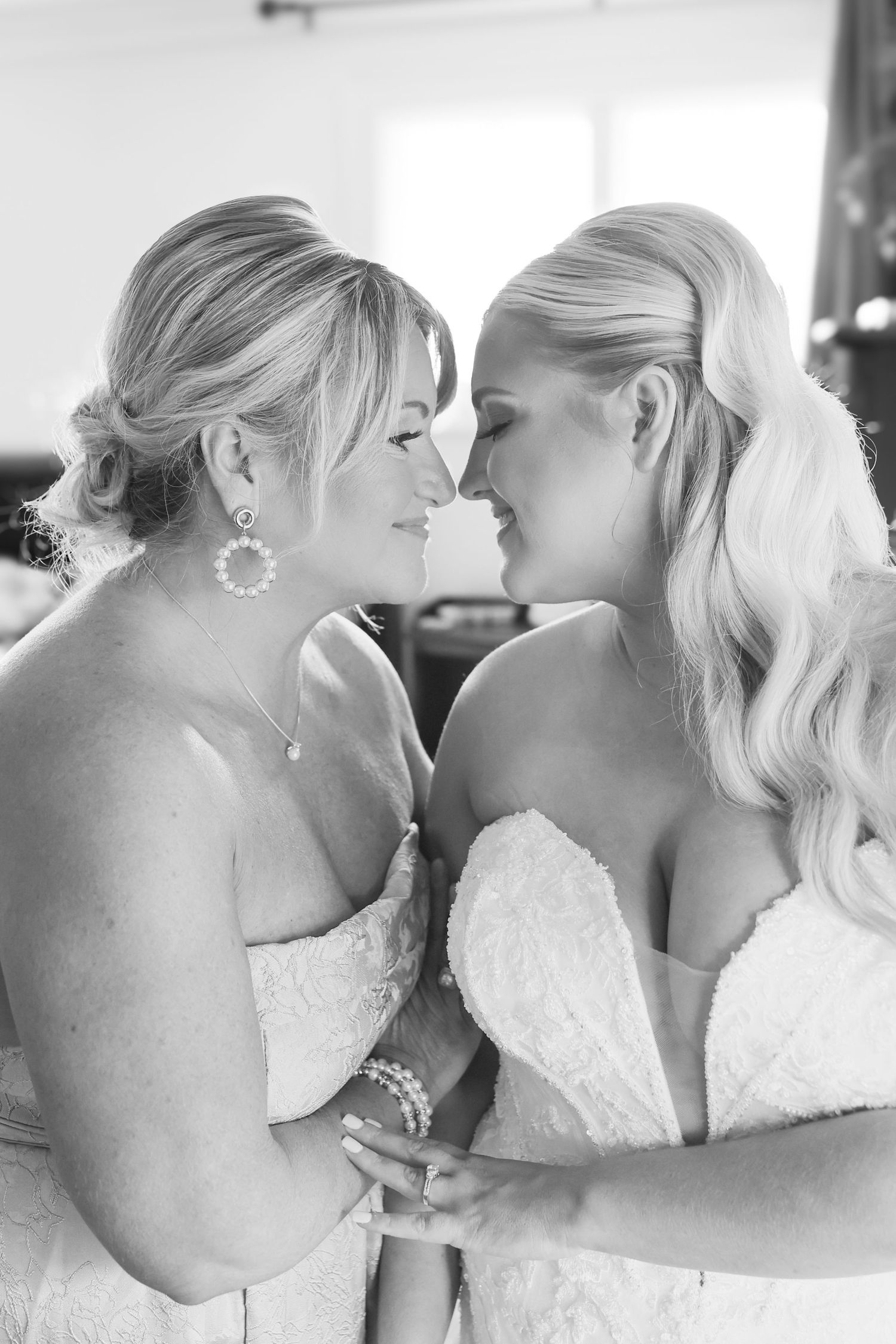 Two brides in white wedding gowns share an intimate moment, touching foreheads and smiling in black and white photo.