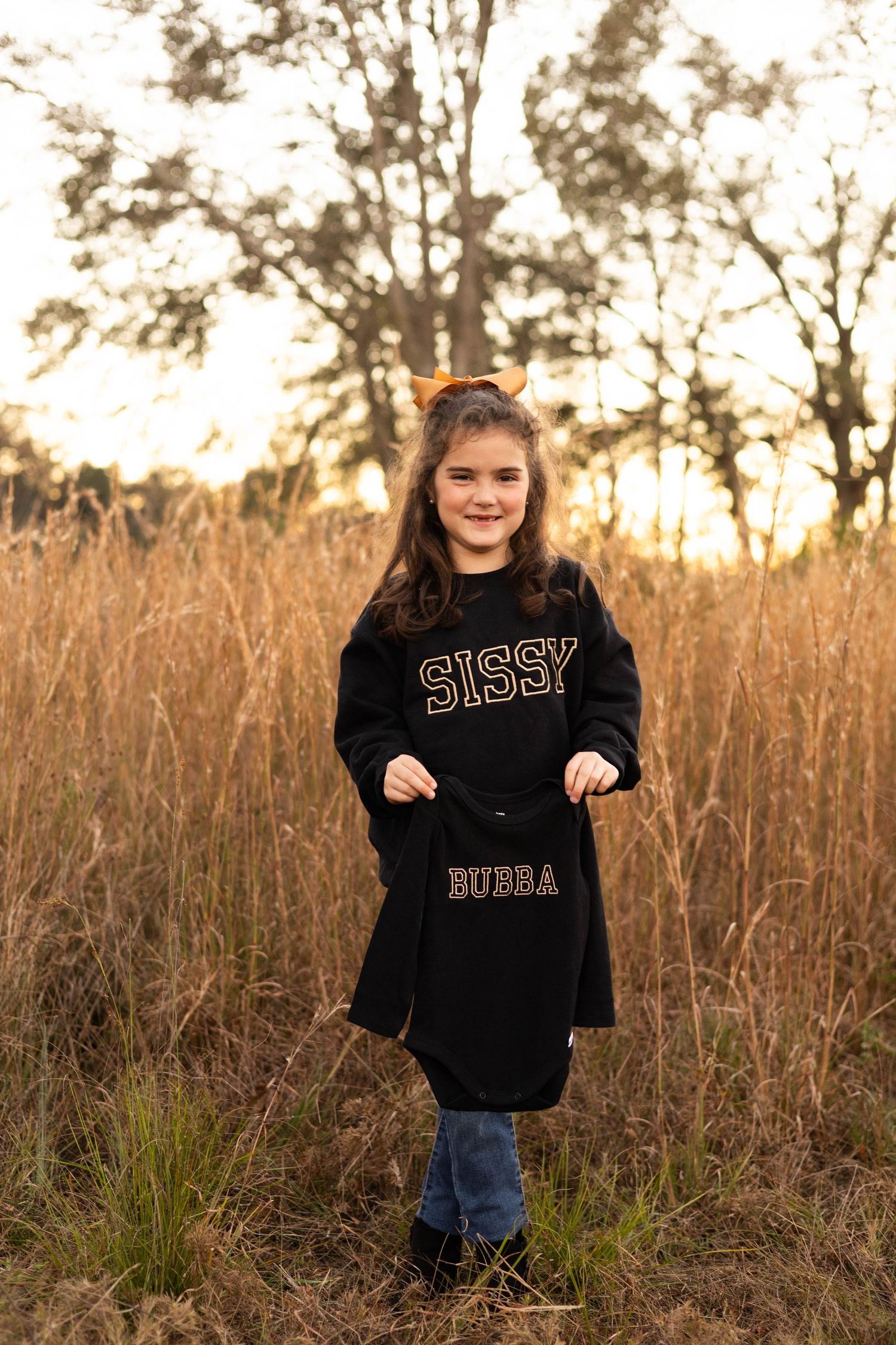 Girl in black 'Sissy' sweatshirt dress holding 'Bubba' onesie, standing in golden field at sunset.