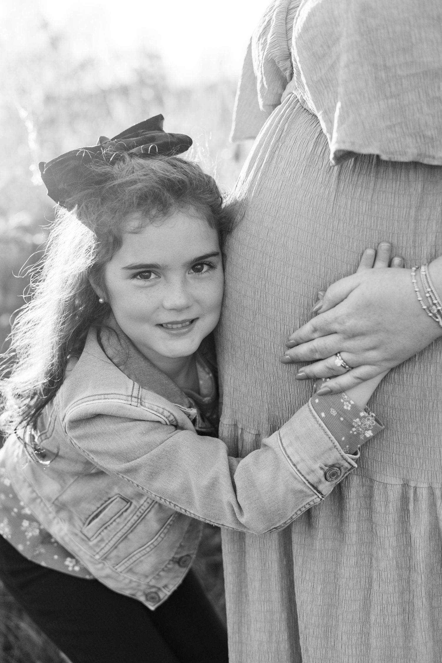 Black and white photo of a young girl hugging a pregnant woman's belly outdoors.
