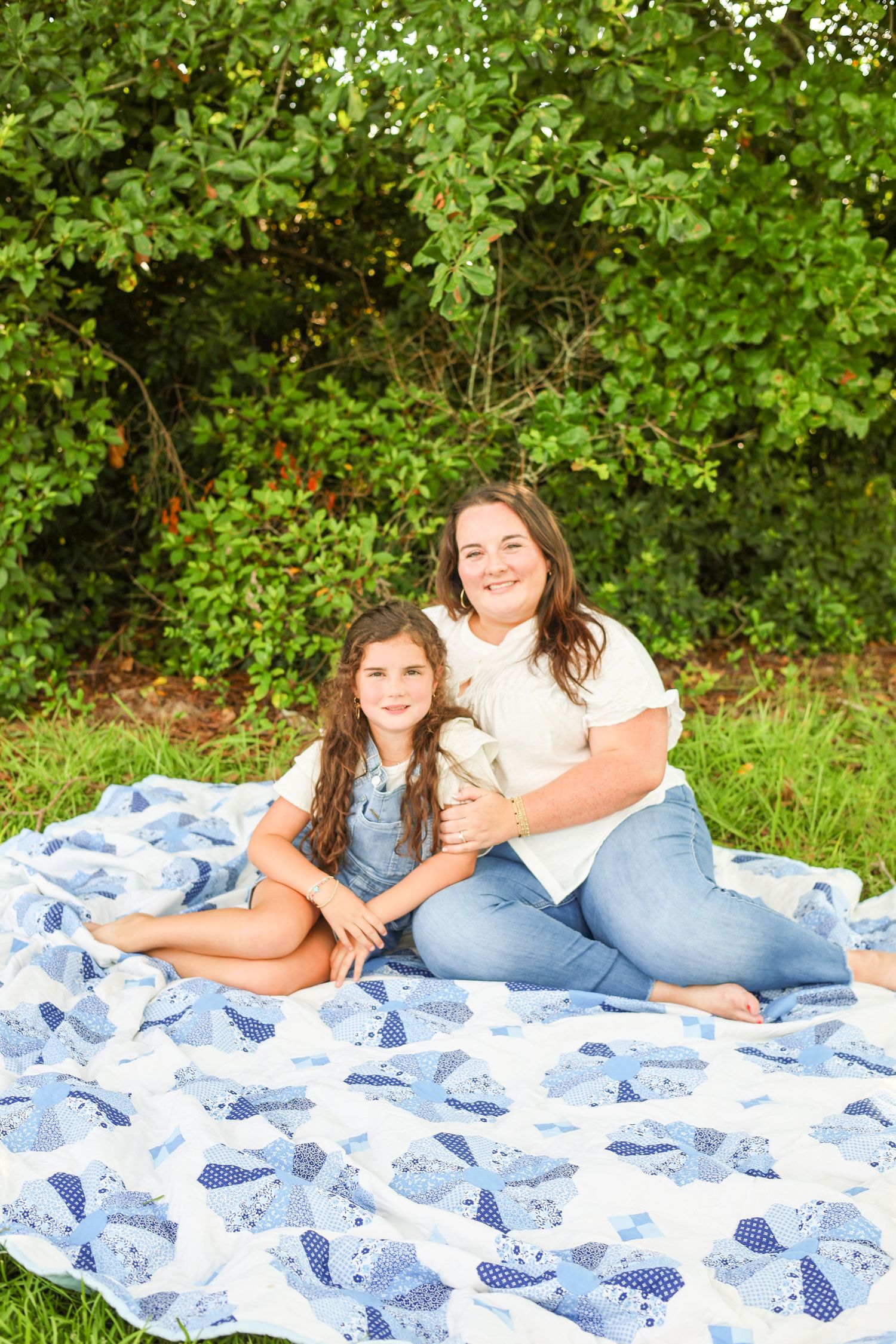 A woman and young girl smile together on a blue patchwork quilt outdoors surrounded by lush green trees.