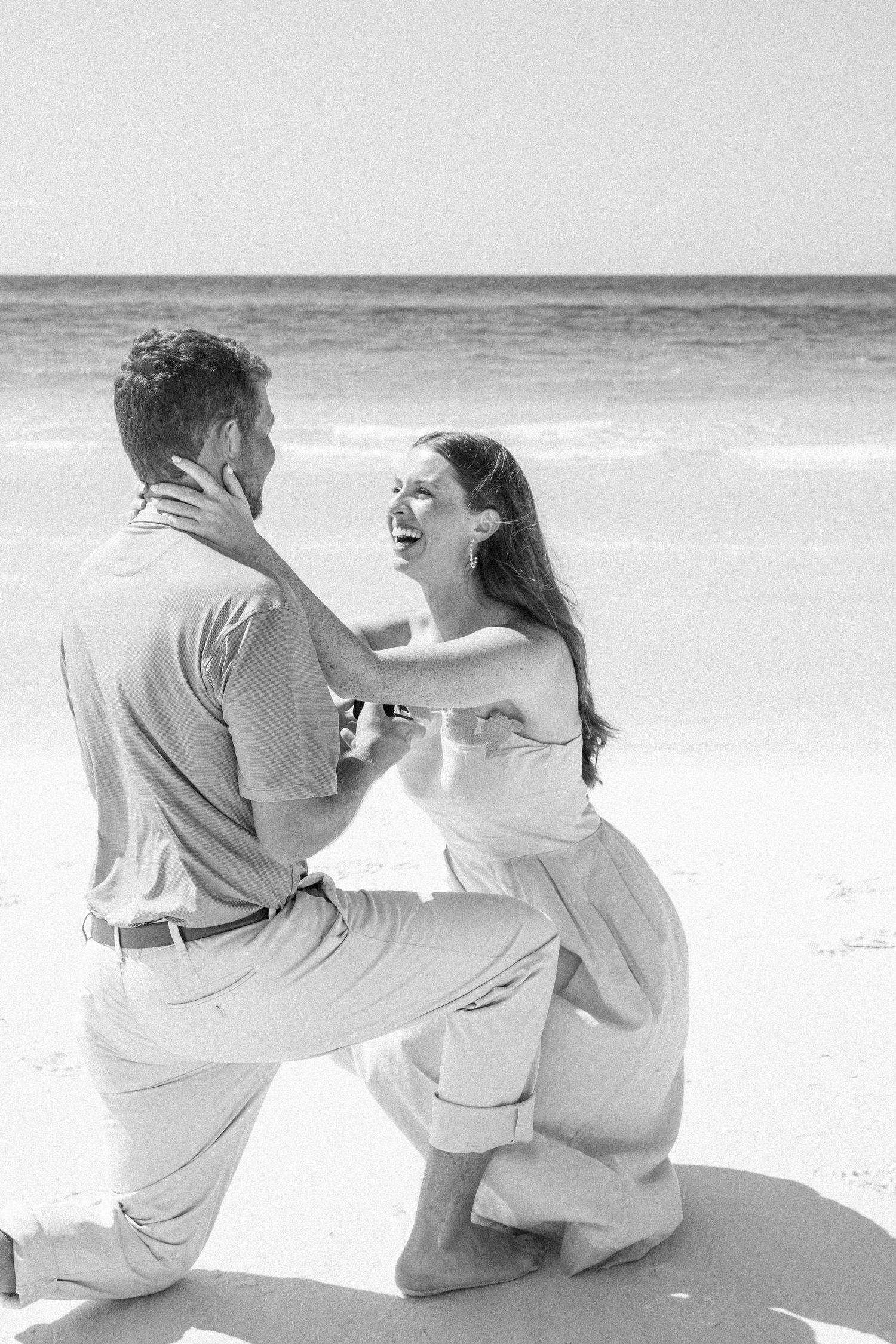 Black and white beach proposal photo of man on one knee as woman laughs joyfully, ocean waves in background.