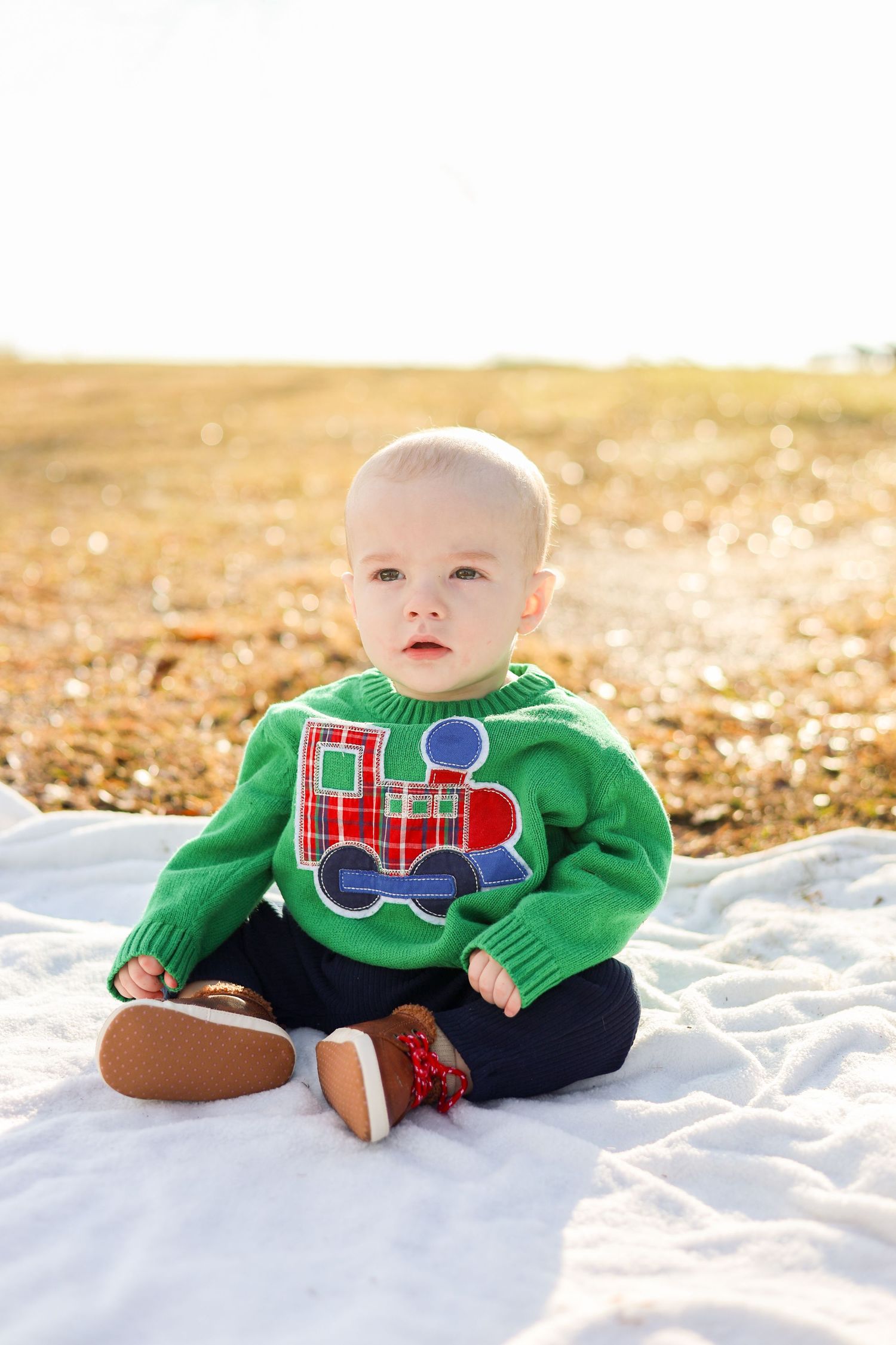 Baby wearing green train sweater lies on white blanket surrounded by autumn leaves in golden light.