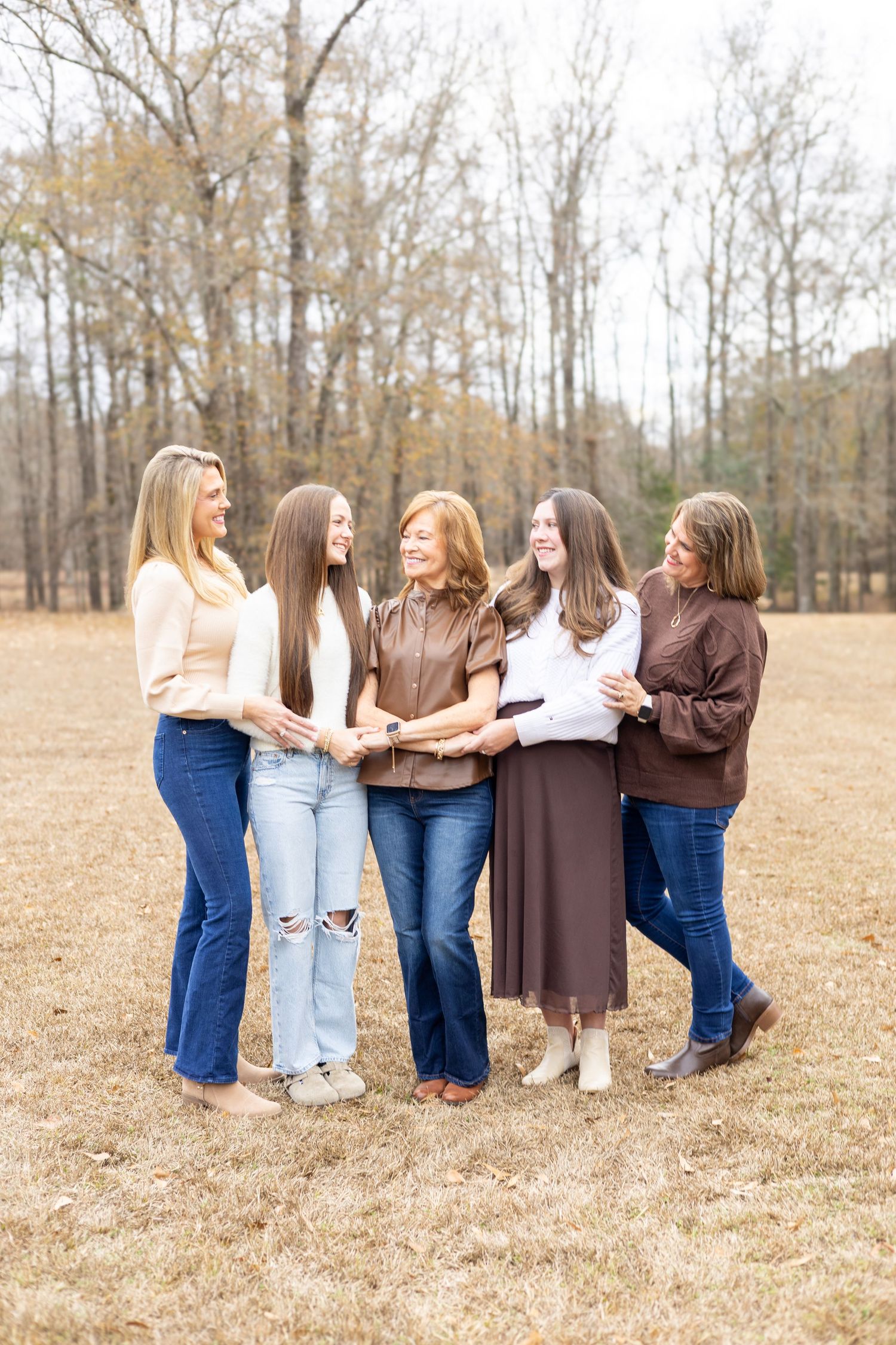 A group wearing jeans and brown tops stand together in a field during autumn with bare trees in the background.