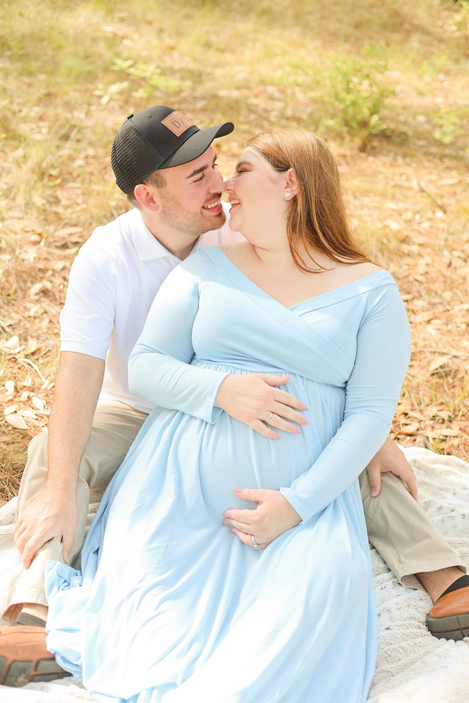 An expectant couple shares intimate moment while sitting on blanket outdoors.