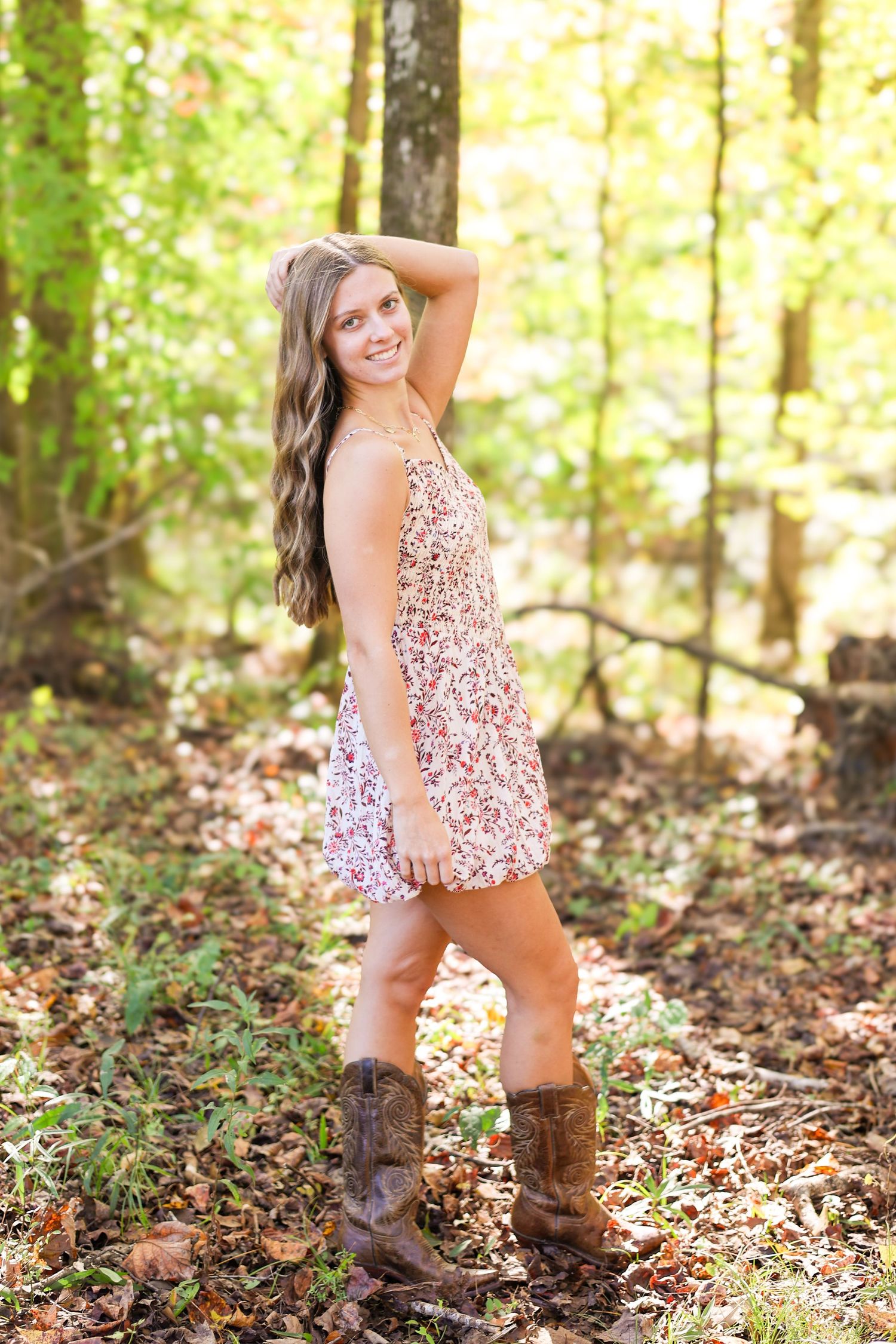 Person in floral dress and brown boots poses playfully in autumn forest.