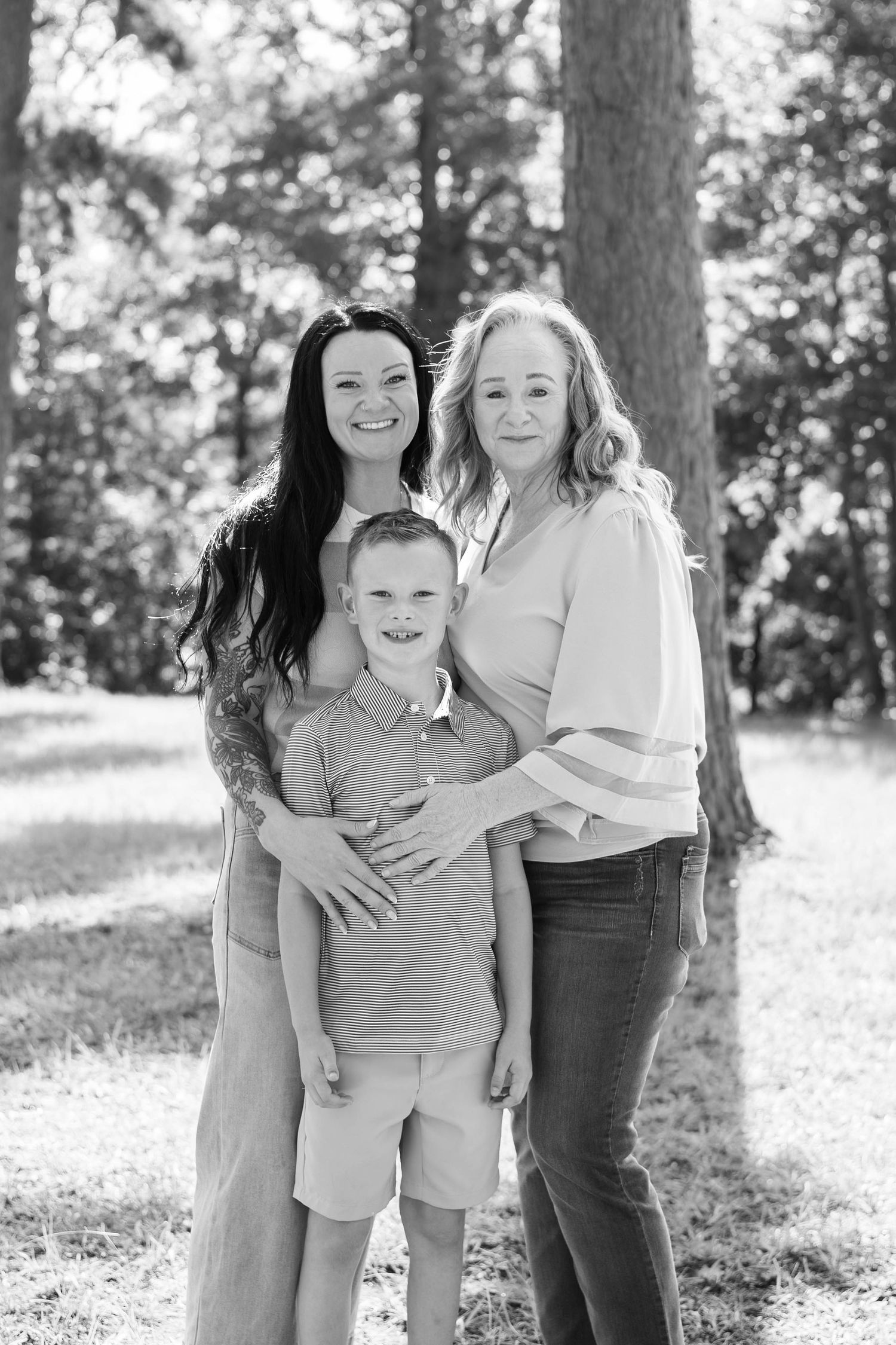 Black and white family portrait of two women and a young boy smiling outdoors in a wooded park setting.
