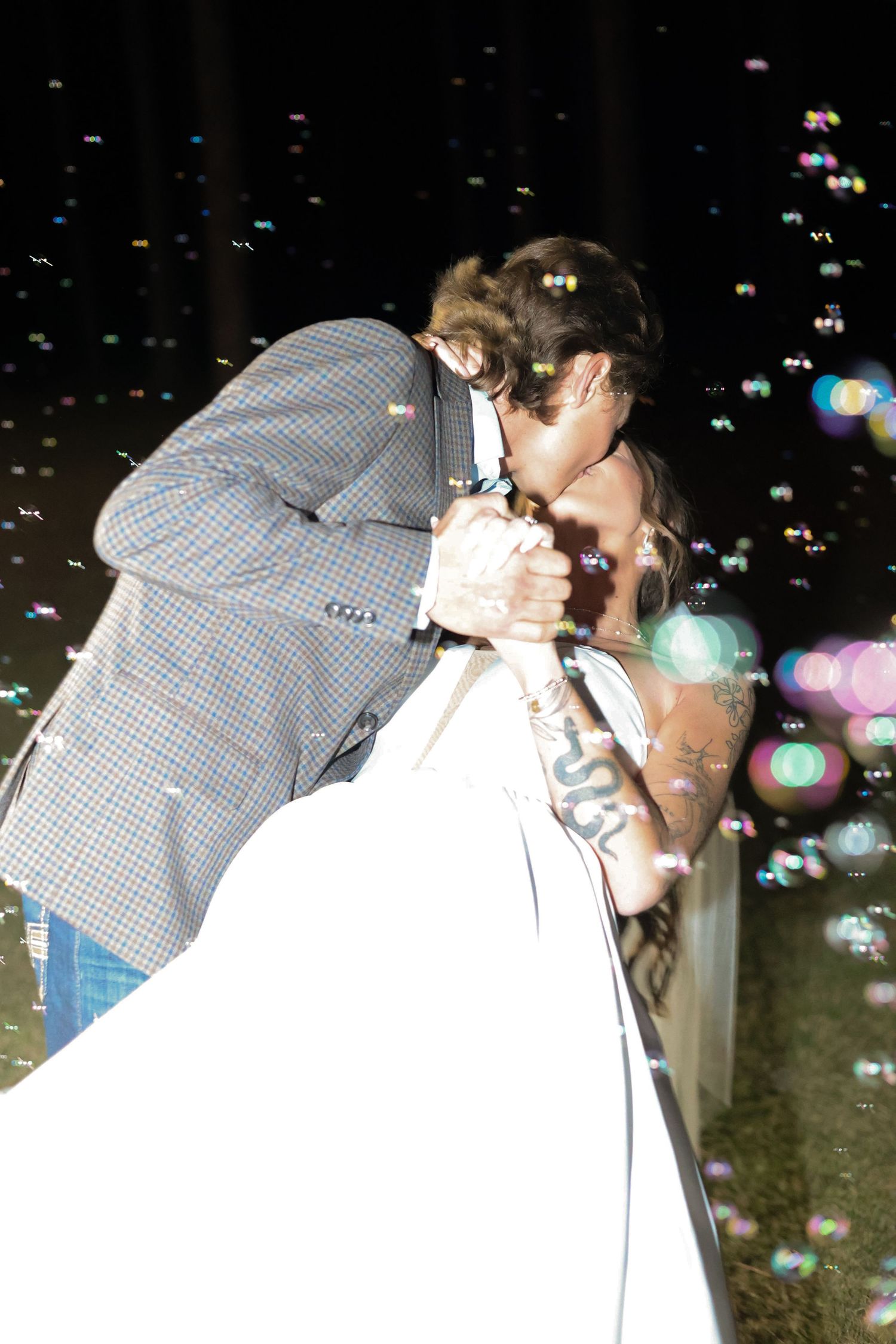 Couple sharing a romantic kiss surrounded by floating bubbles at night, bride in white dress and groom in plaid jacket.