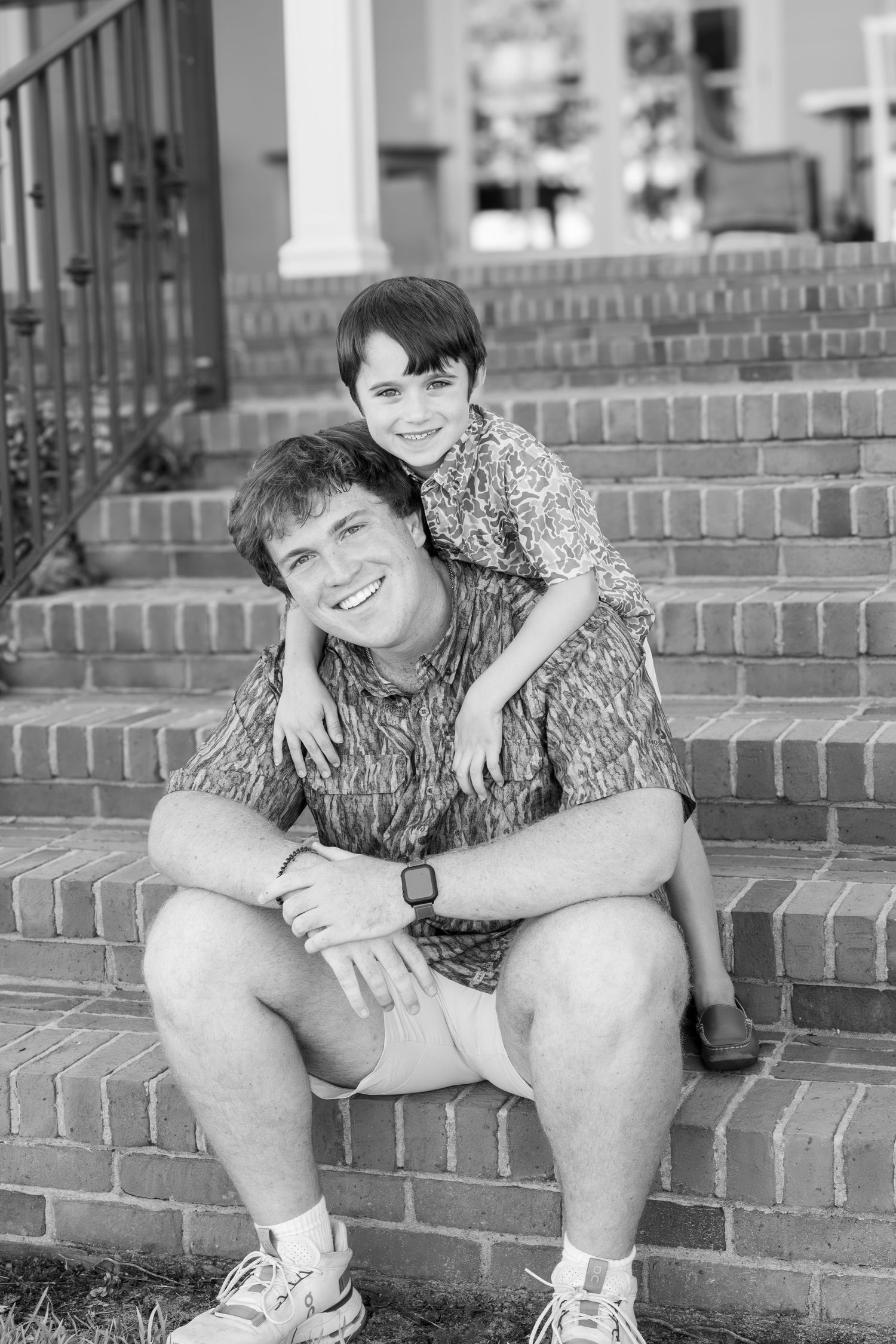Black and white photo of a smiling man sitting on brick steps with a young boy leaning on his shoulders.