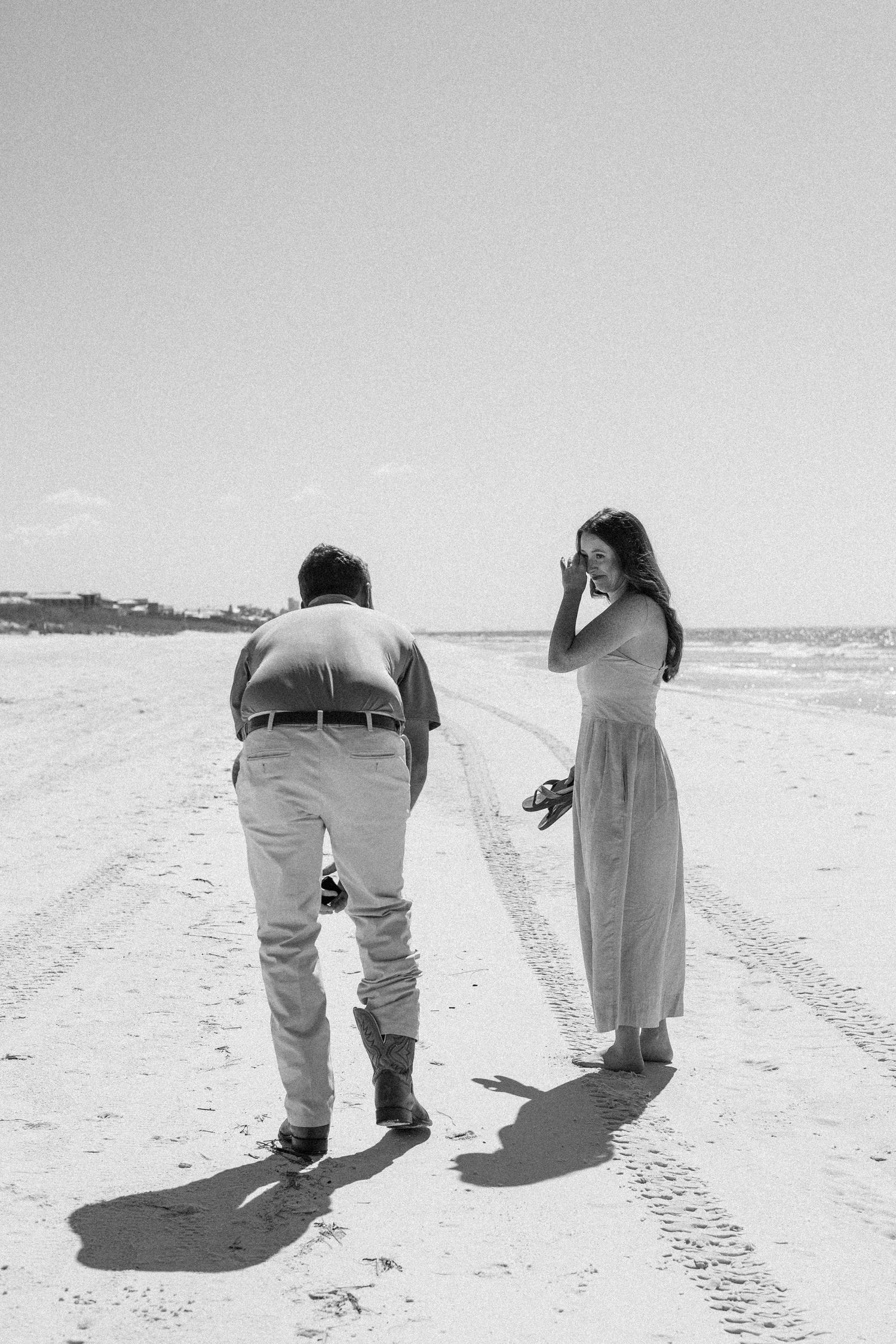 Black and white photo of a couple on a sandy beach, man bending down while woman in maxi dress holds her shoes.