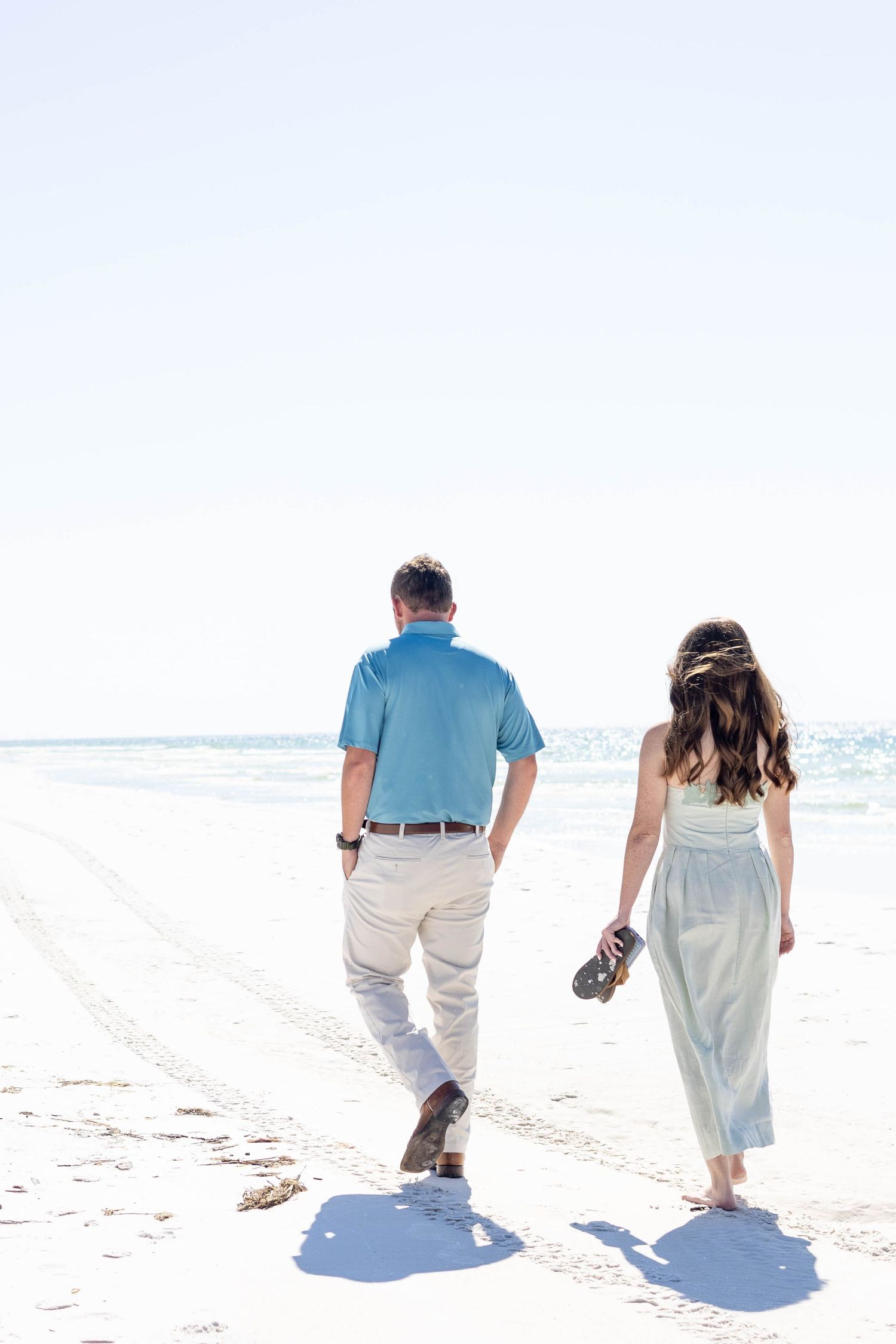 A couple strolls along a pristine white sandy beach carrying their shoes.