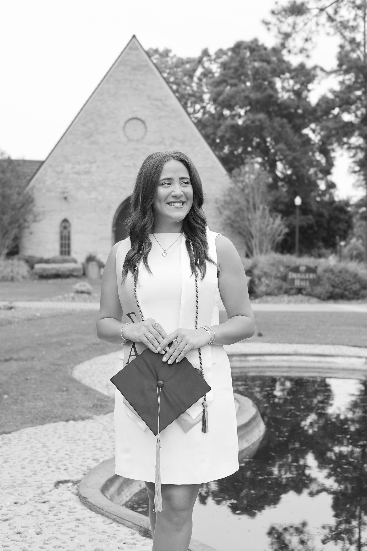 Graduate holds diploma and poses near stone chapel with reflection pool.