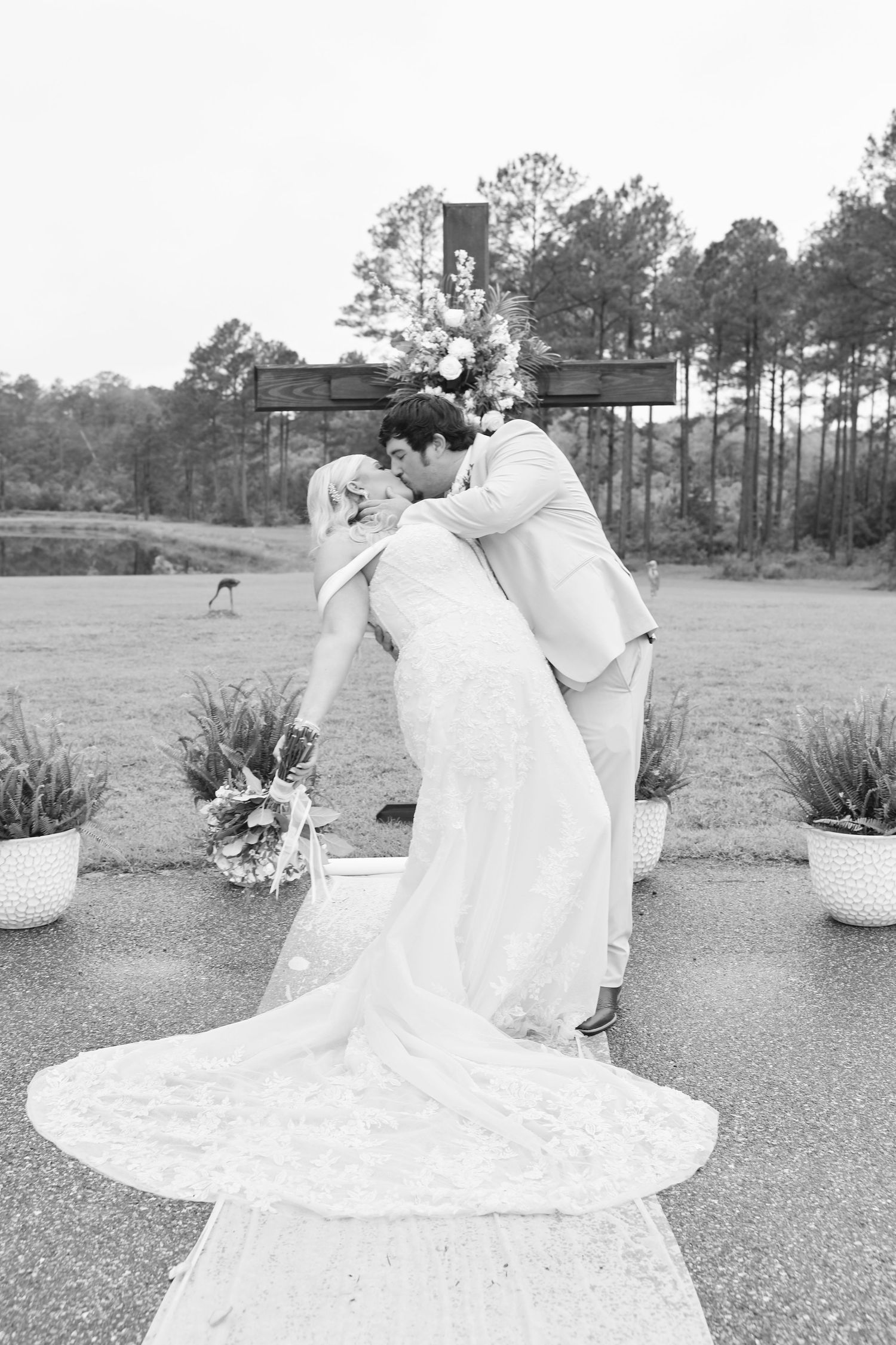 Bride and groom share a romantic dip kiss at outdoor wedding ceremony with floral cross backdrop.