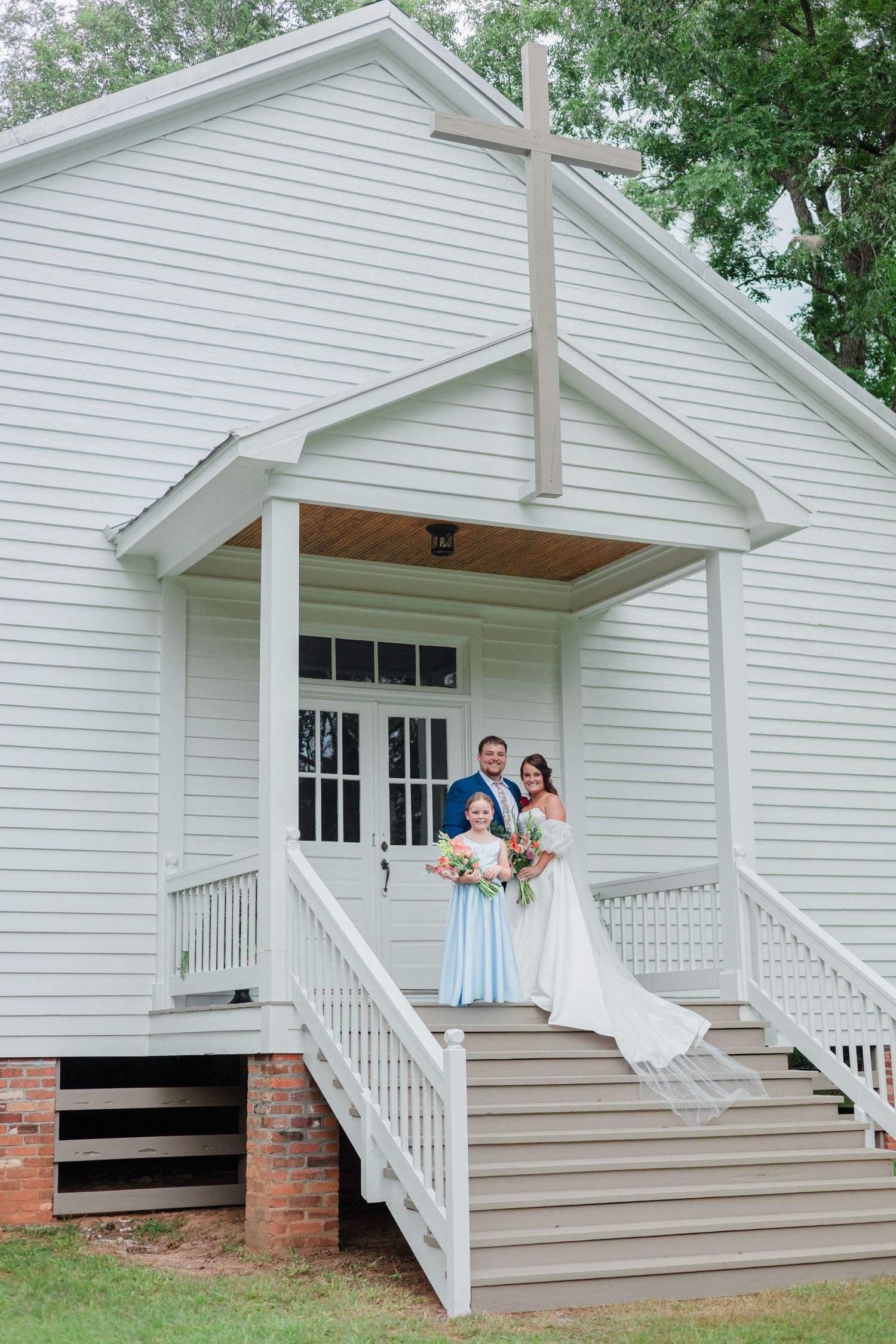 Bride and groom pose with bridesmaid on steps of charming white chapel with cross at outdoor wedding ceremony.