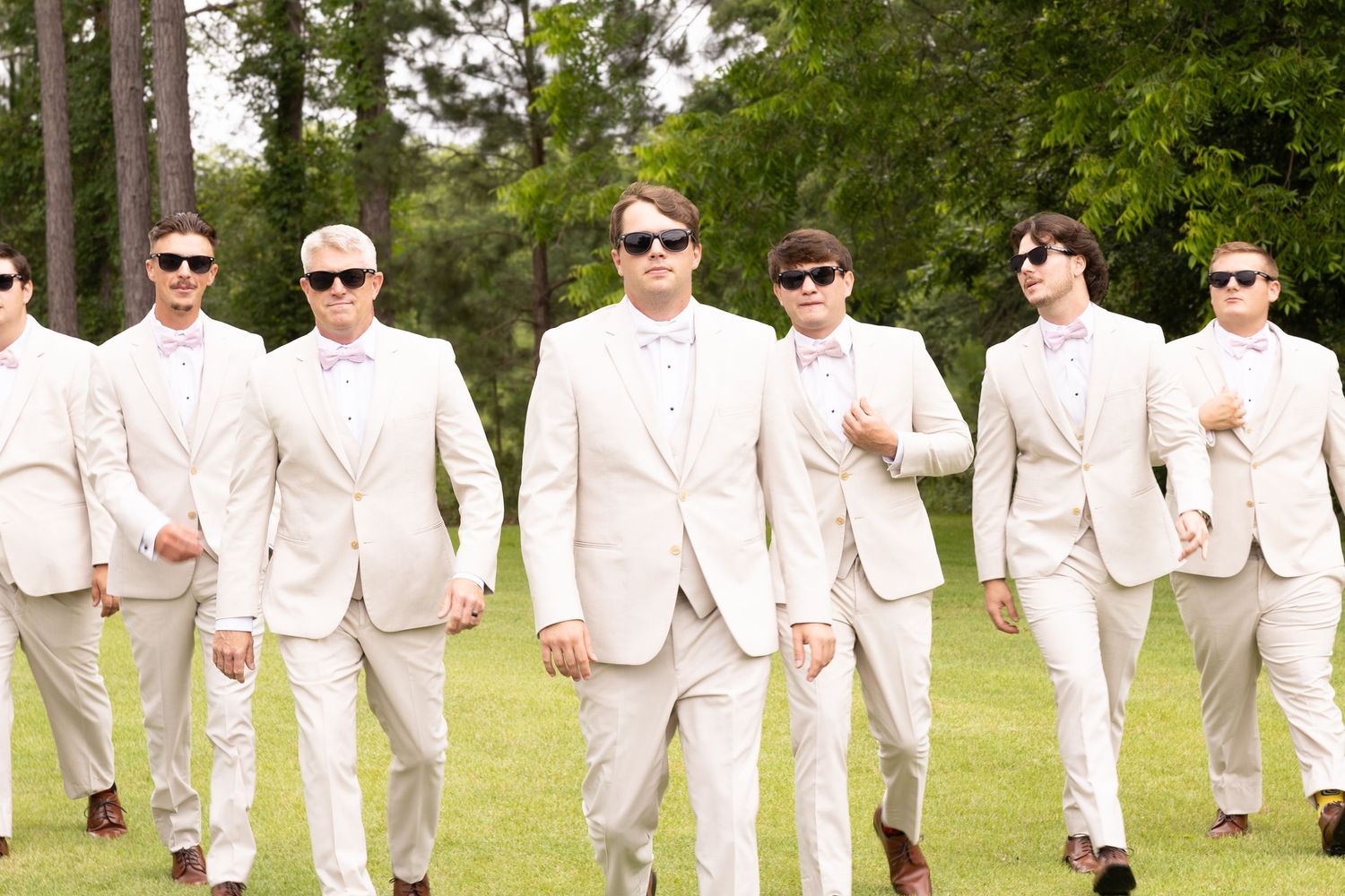 Groomsmen in matching cream suits and sunglasses walking outdoors on green grass at a wedding.