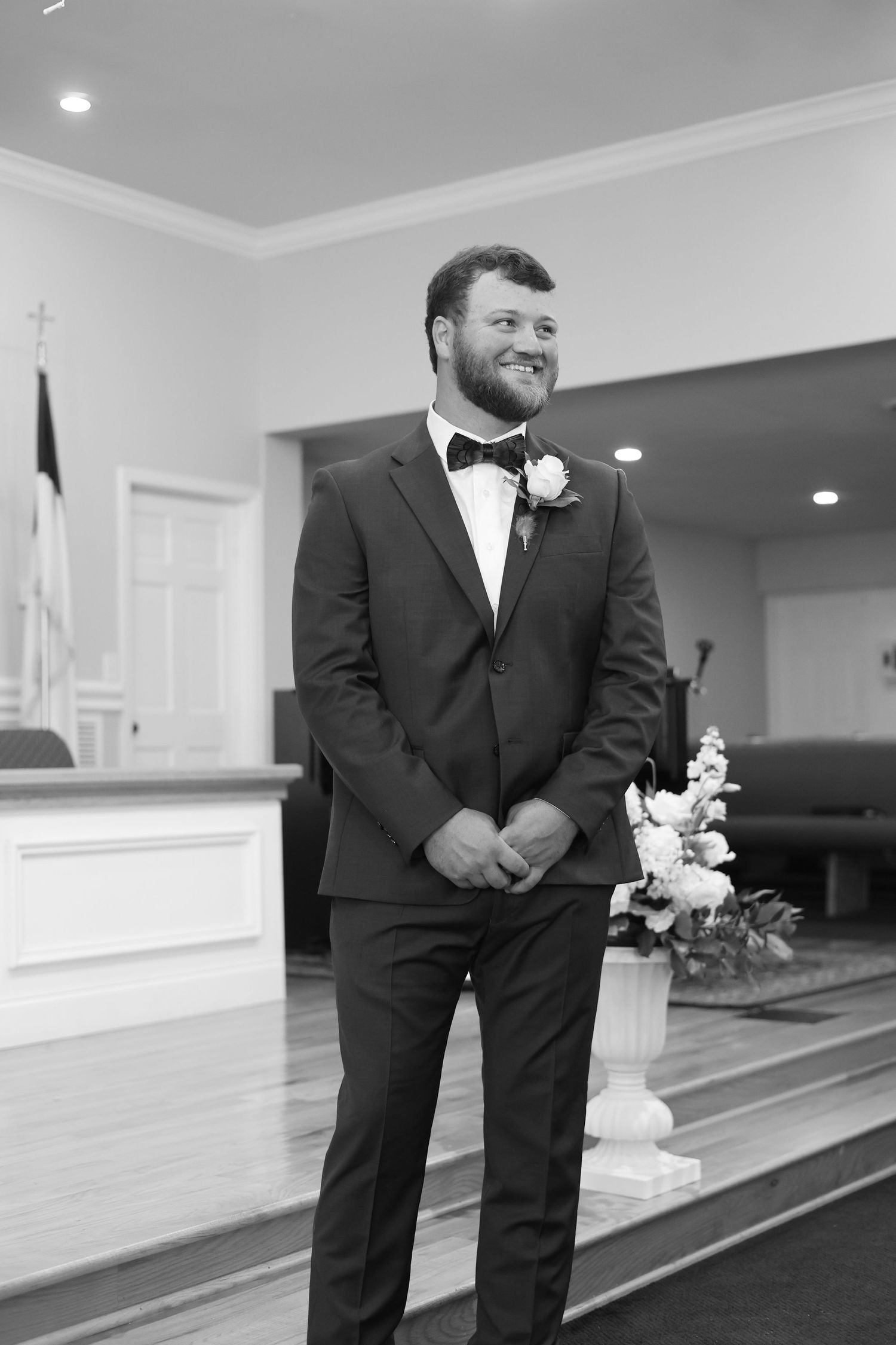 Smiling groom in tuxedo with boutonniere stands at church altar awaiting bride in black and white wedding photo.