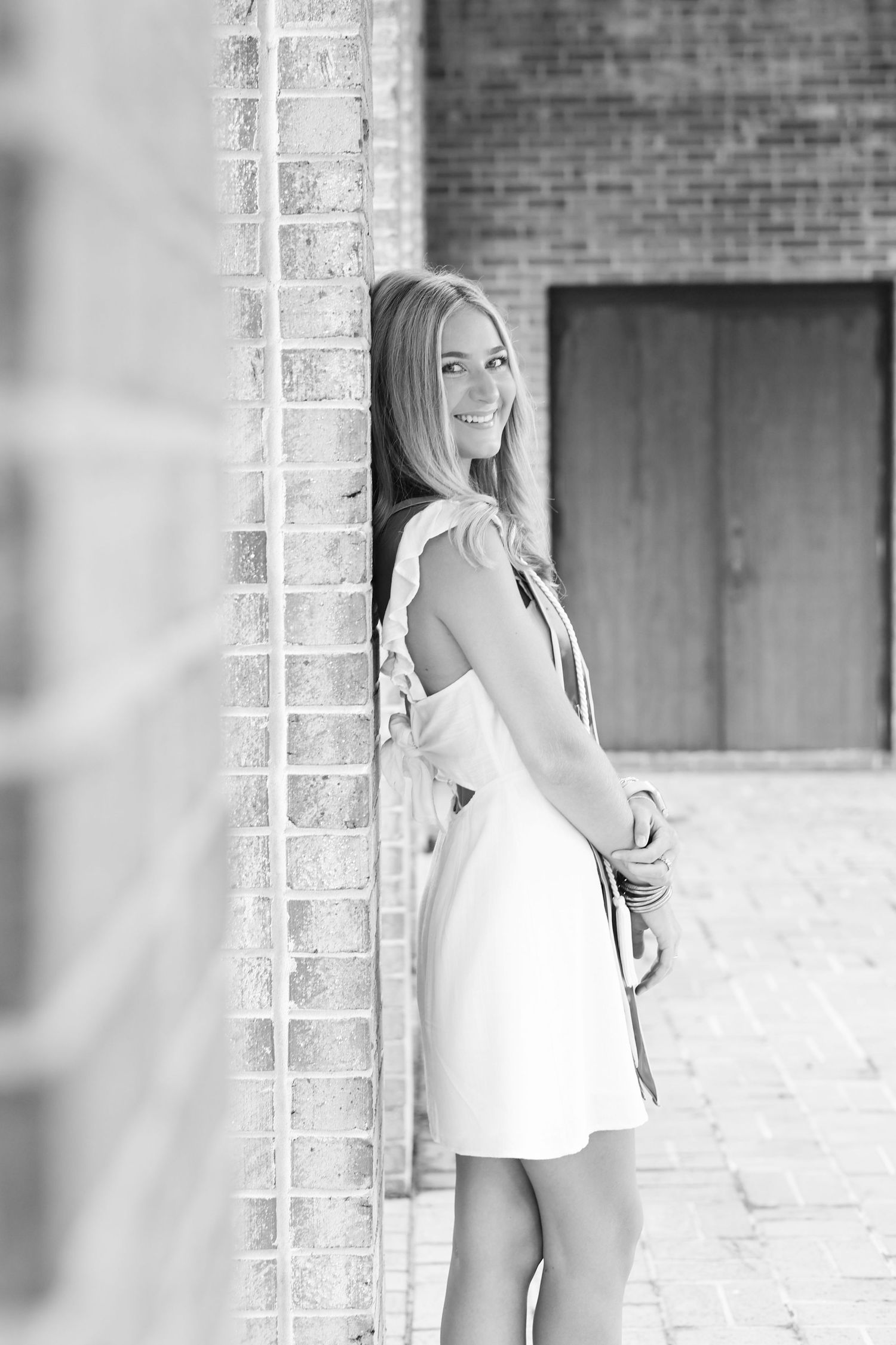 Black and white portrait of graduate in white dress near brick wall.