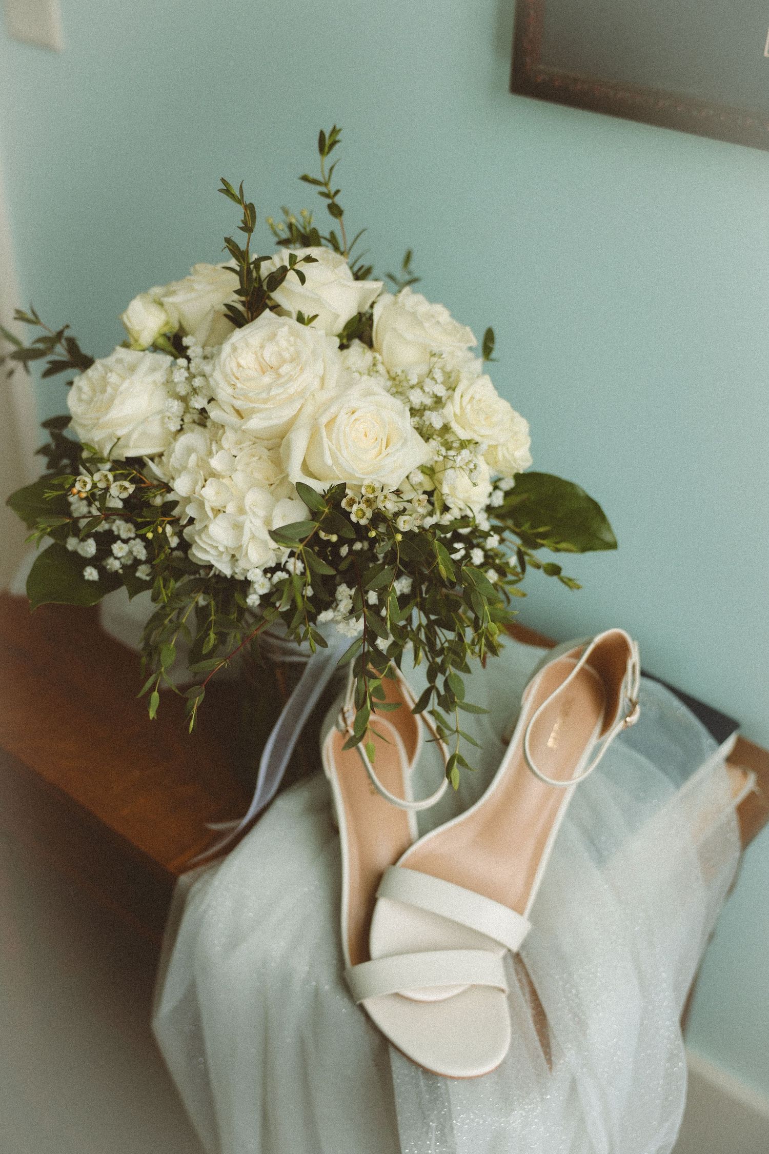 Elegant bridal bouquet of white roses and greenery beside white strappy wedding heels on a wooden surface.