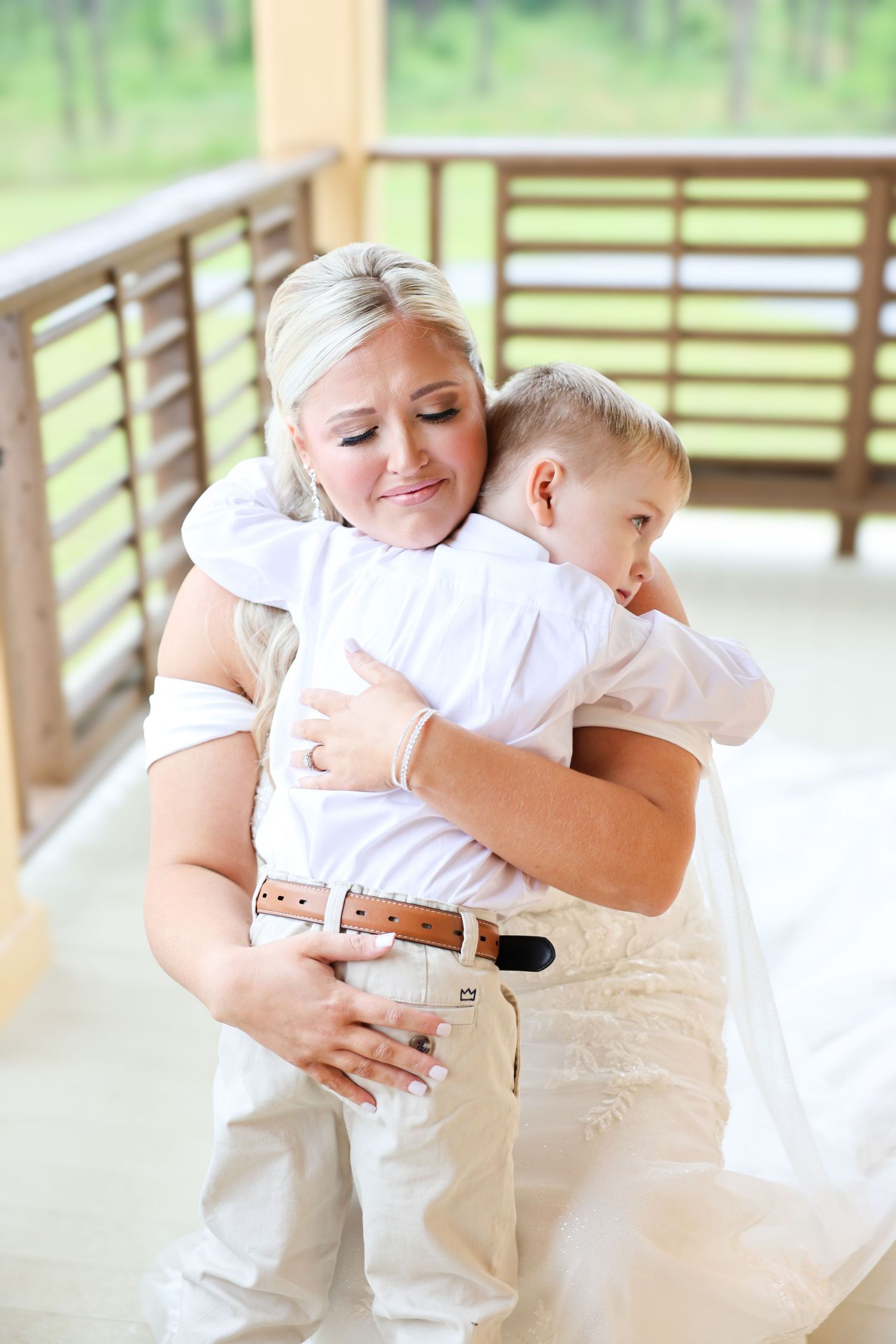 Bride in wedding dress hugging young boy in white shirt and khaki pants on a wooden porch.