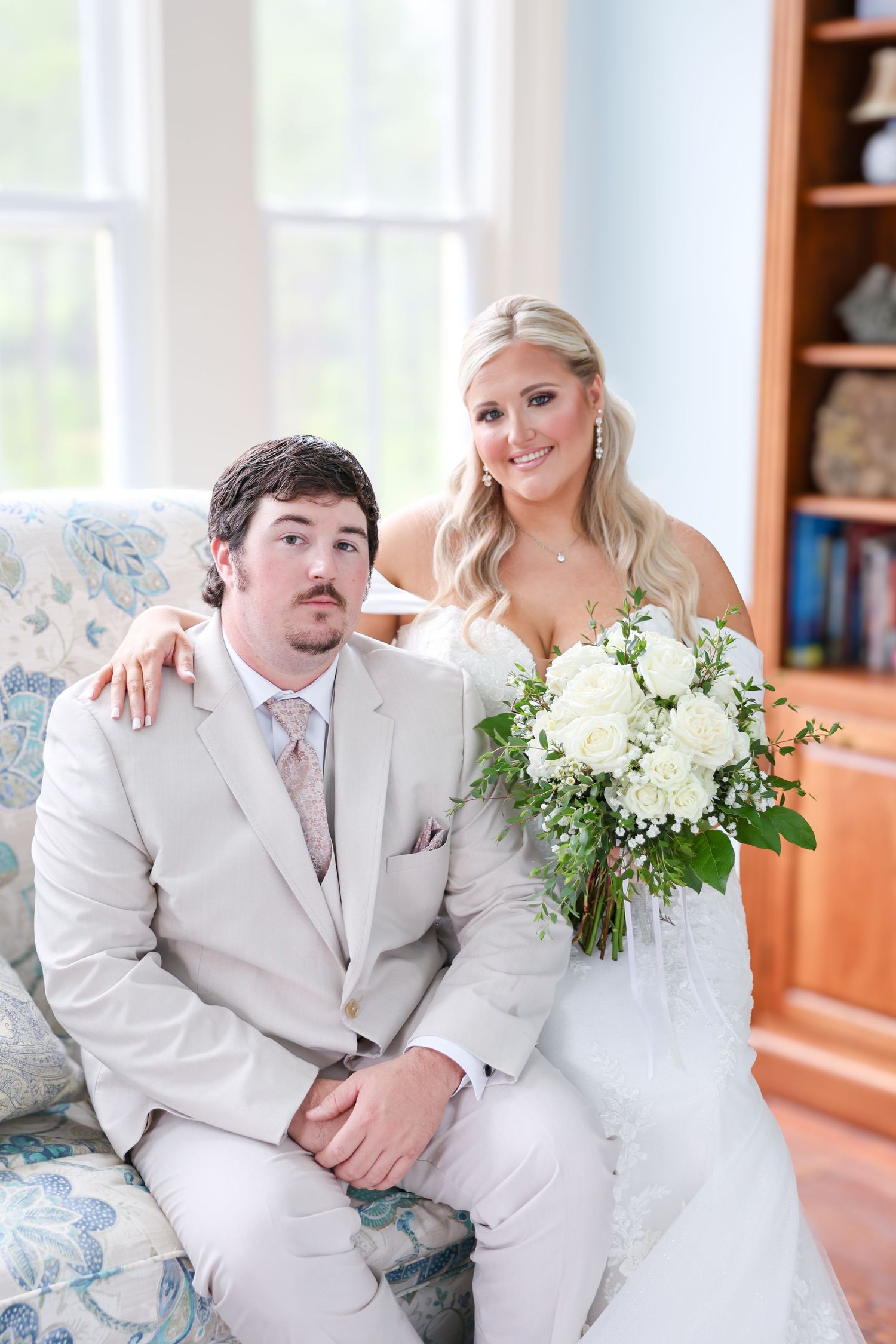 Smiling bride in white lace gown holding white bouquet poses with groom in tan suit on floral sofa.