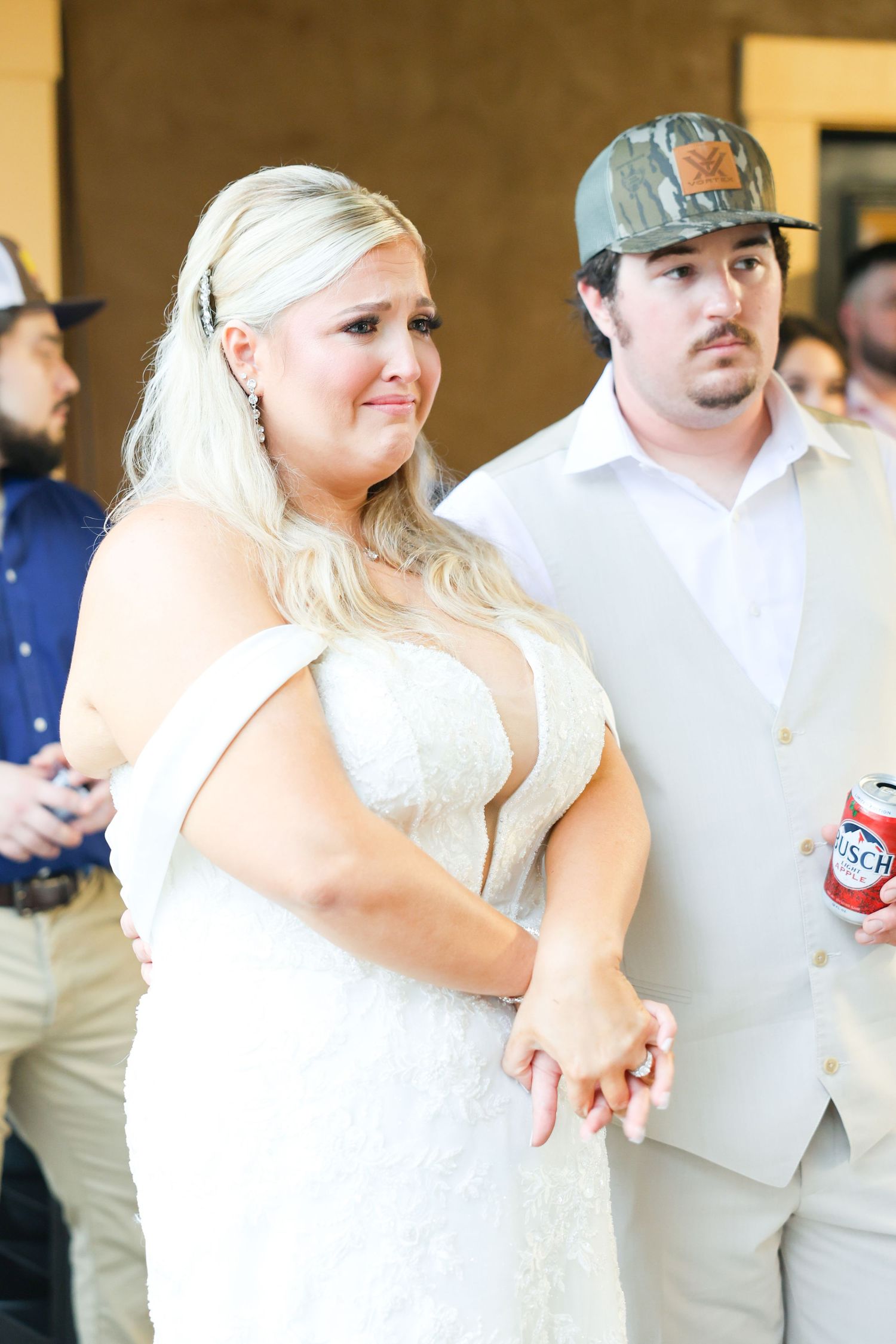 Emotional bride in lace gown holds hands with groom in camo hat at rustic wedding reception.