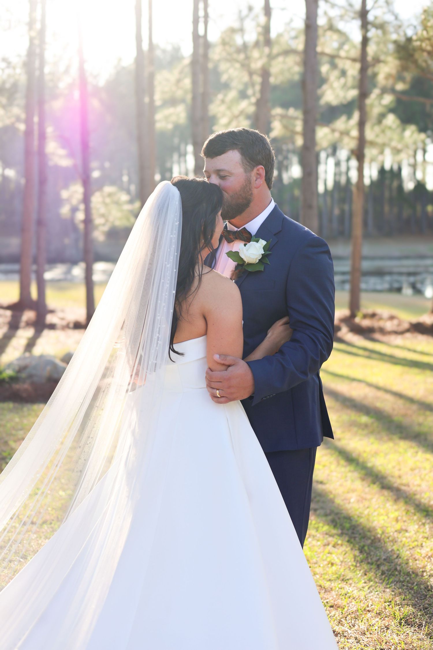 Bride and groom sharing a romantic kiss outdoors among tall pine trees with golden sunlight.