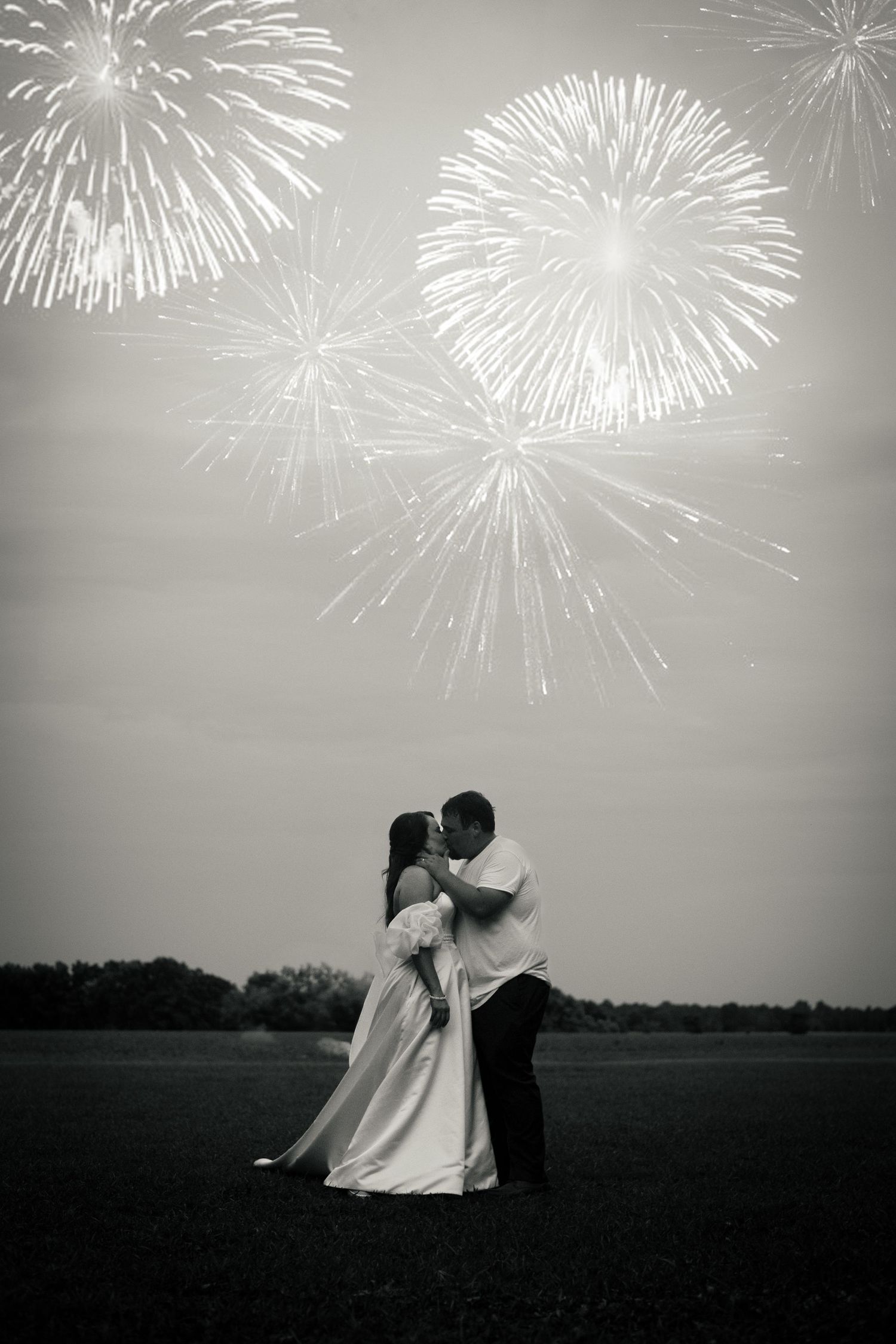 Bride and groom share a romantic kiss in a field with spectacular fireworks display overhead in black and white photo.