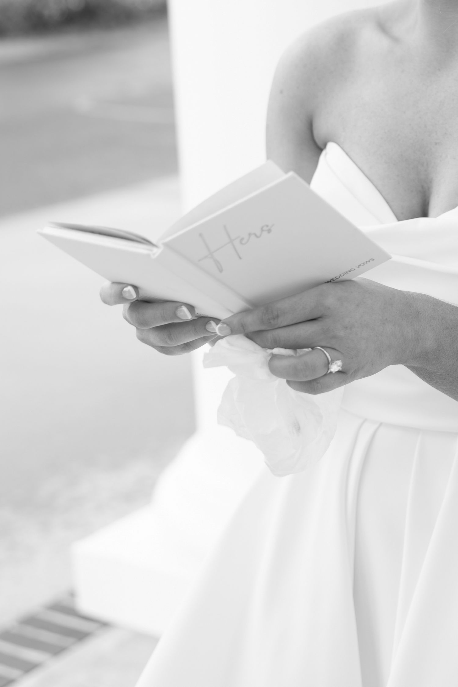 Bride in white gown reads wedding vows from a small book, engagement ring visible on her hand.