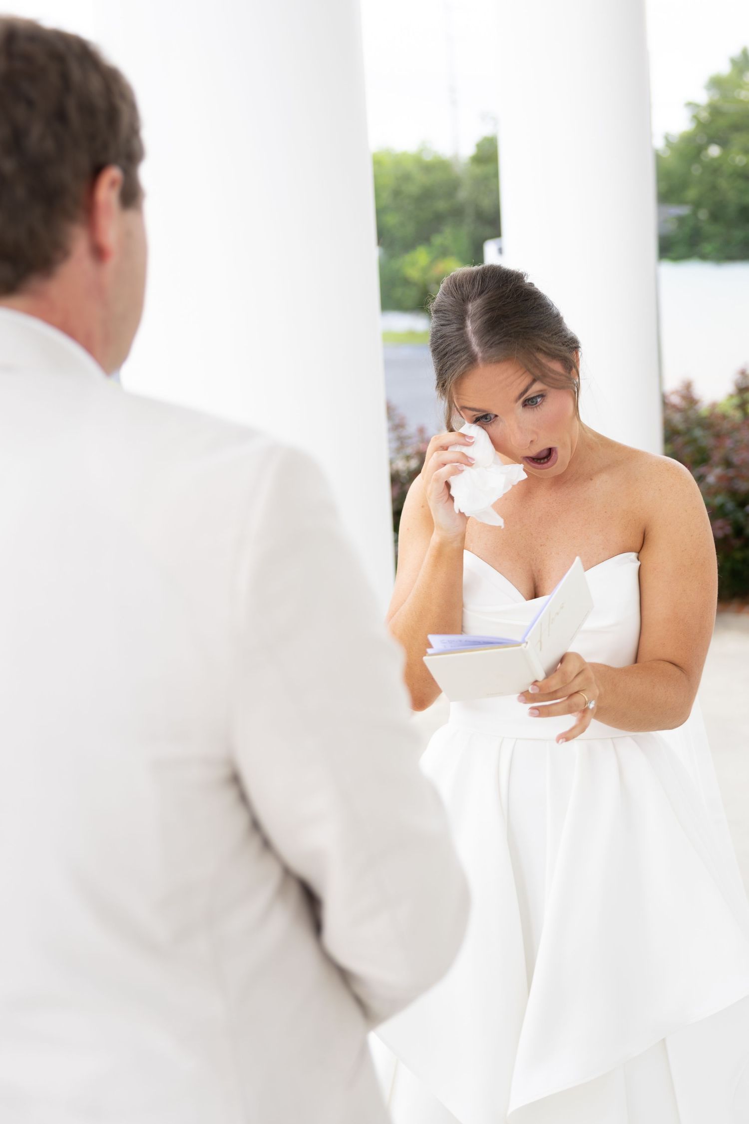 Emotional bride in white strapless dress wiping tears while reading vows during outdoor wedding ceremony.
