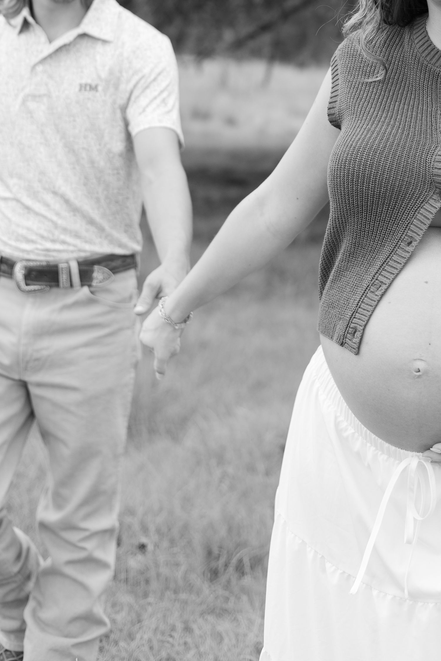 Black and white maternity photo of couple holding hands outdoors, showing pregnant belly in knit top and white skirt.