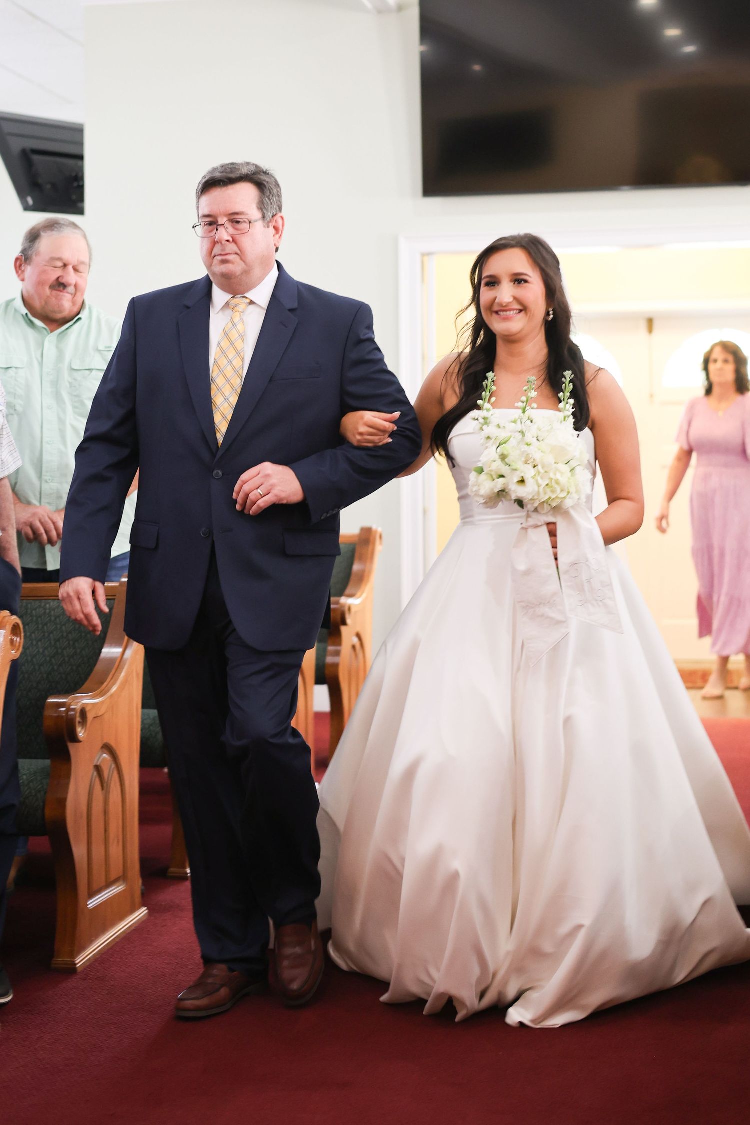 A wedding processional shows a bride in a white gown walking down a red-carpeted church aisle.
