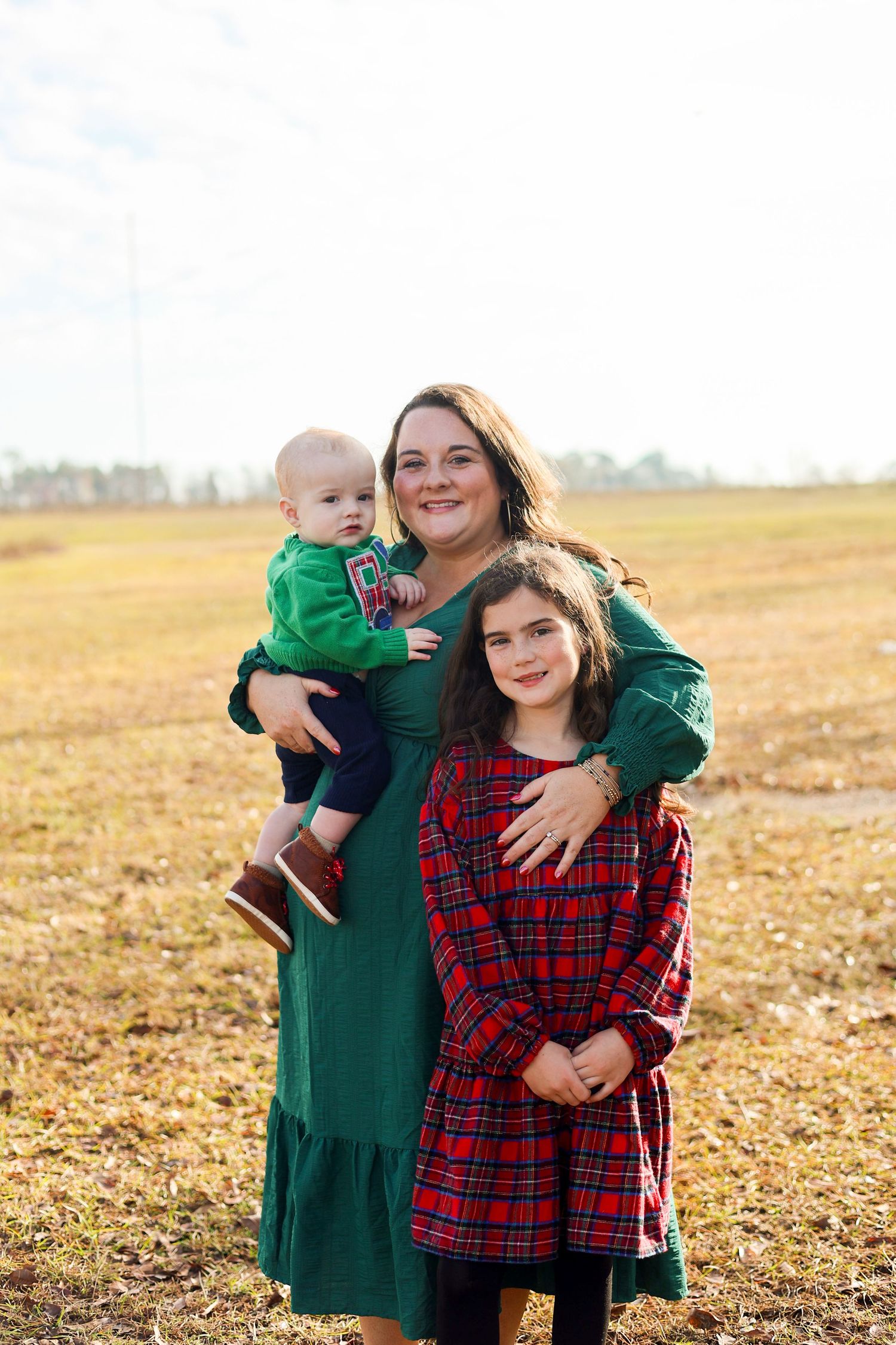 A family wearing coordinating green and red plaid dresses poses together in a golden autumn field at sunset.
