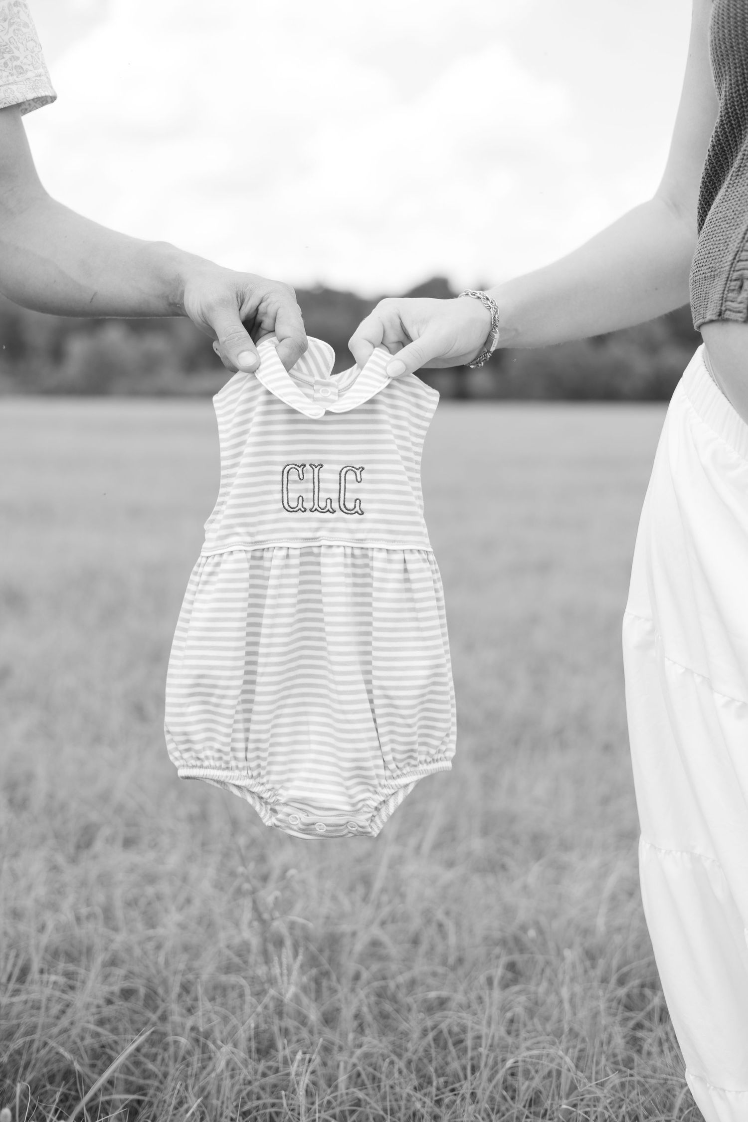 Two people holding a monogrammed striped baby romper outdoors in a field, black and white pregnancy announcement photo.