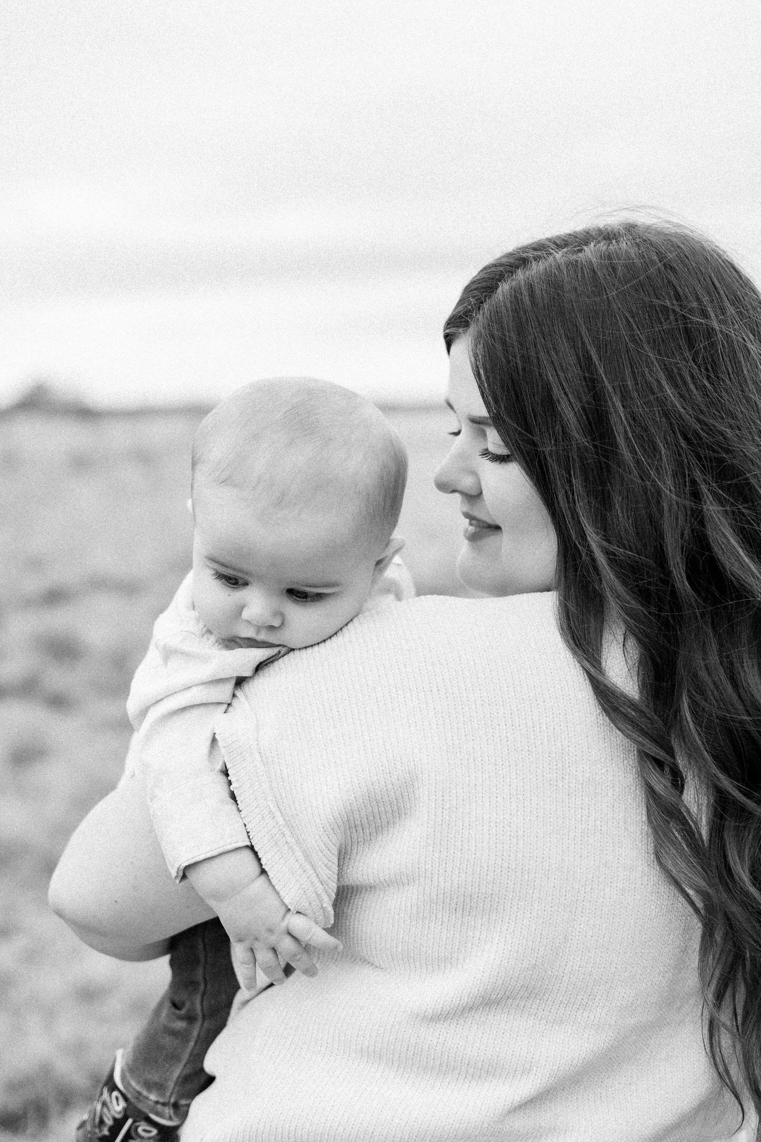 Black and white photo of a mother with long dark hair holding her baby outdoors in a field.