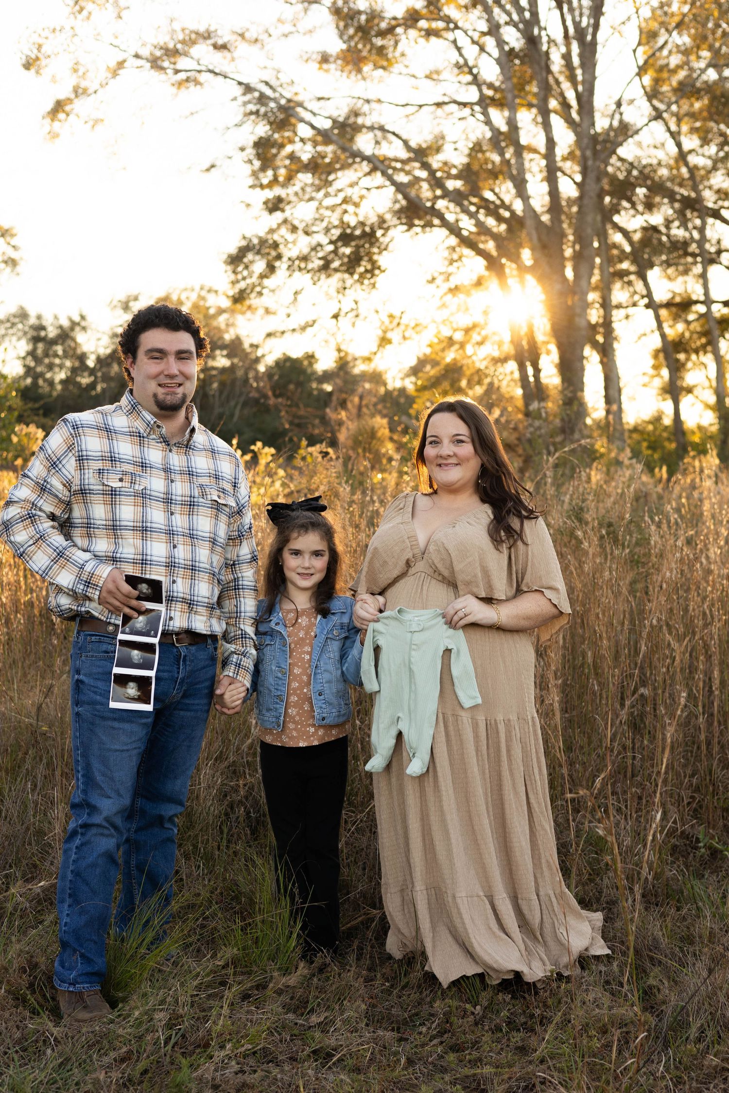 Family of three posing at golden hour in a field, holding ultrasound photos and baby clothes, announcing a pregnancy.